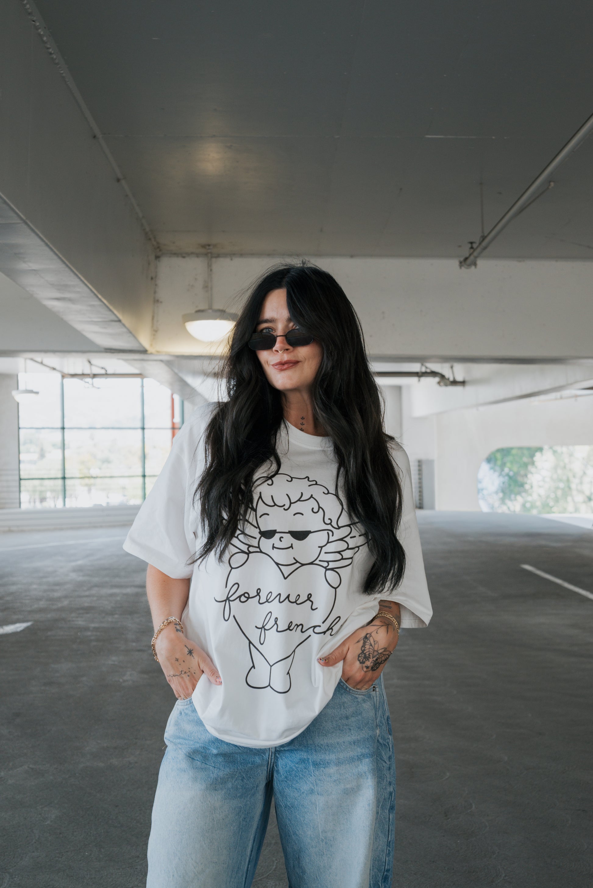 A woman with long dark hair and sunglasses stands in a parking garage, wearing the forever french baby Adult Cotton Tee | Big Baby—a unisex cotton t-shirt featuring an angel graphic and the words “forever friend”—with her hands in her pockets and light blue jeans, perfect for everyday wear.
