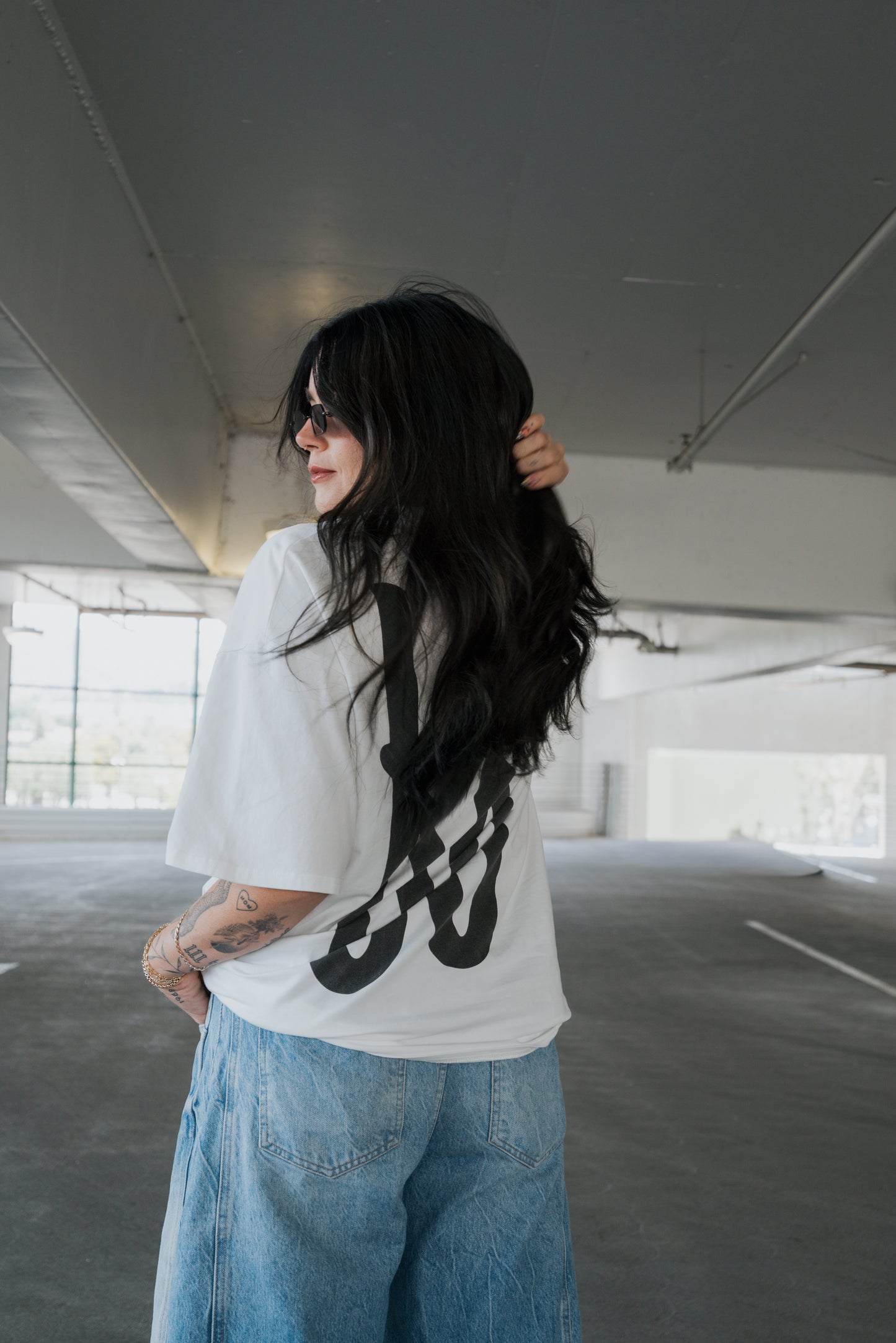 A person with long dark hair stands in a parking garage, wearing the forever french baby Adult Cotton Tee | Big Baby featuring a bold black design, paired with loose blue jeans and glancing over their shoulder—a perfect look for everyday wear.