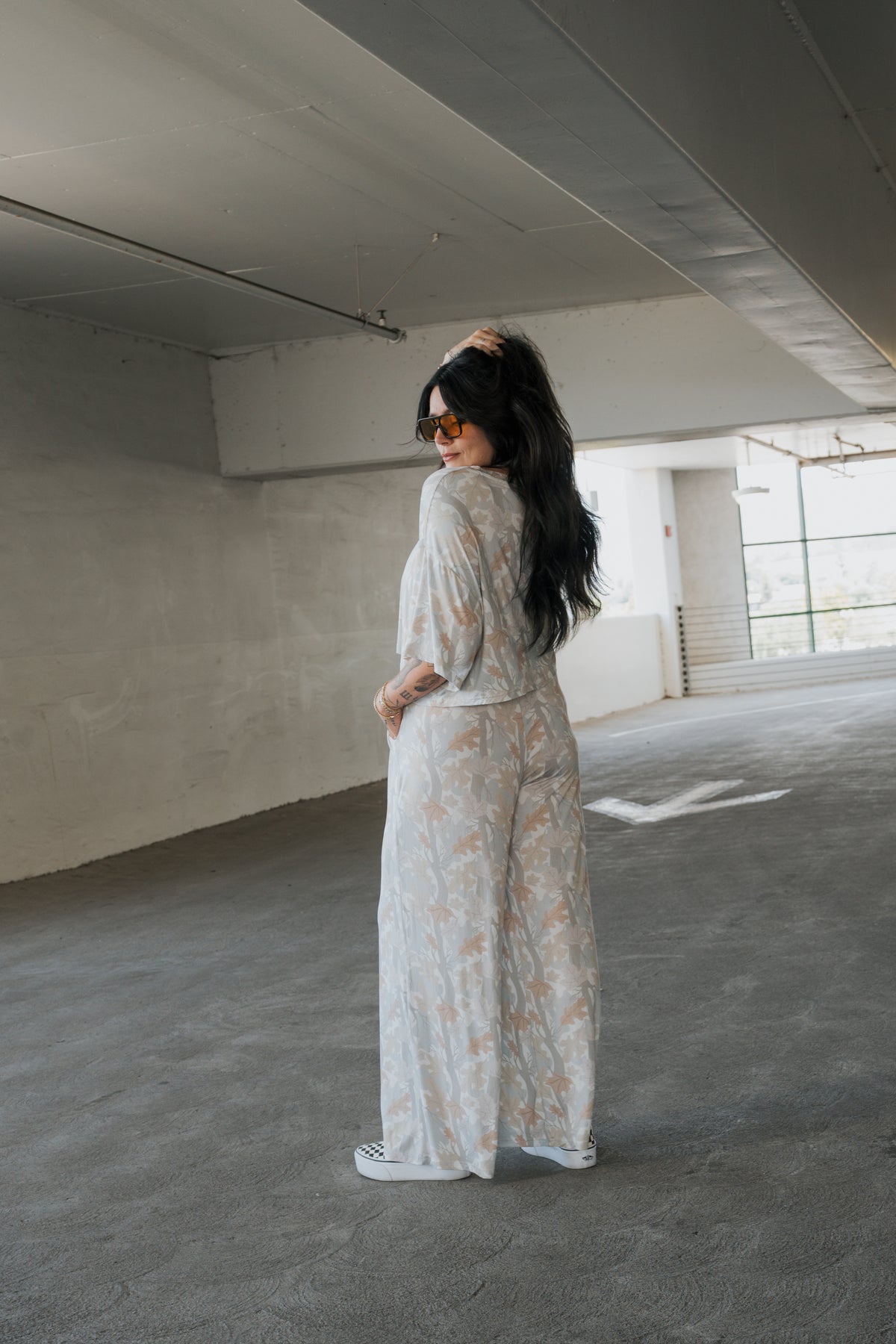A woman with long dark hair, wearing sunglasses and the forever french baby Women’s Bamboo Crop Tee Lounge Set in Woodland, stands in a parking garage, turning slightly to look back over her shoulder.