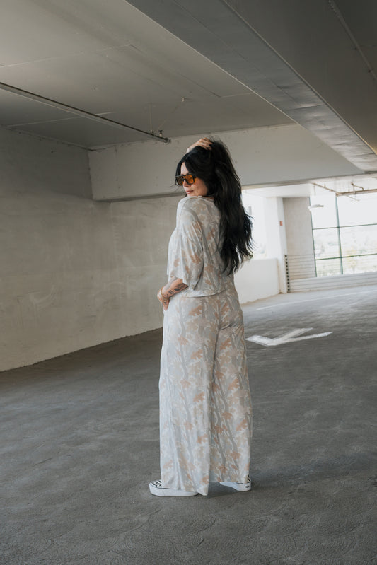 A woman with long dark hair, wearing sunglasses and the forever french baby Women’s Bamboo Crop Tee Lounge Set in Woodland, stands in a parking garage, turning slightly to look back over her shoulder.