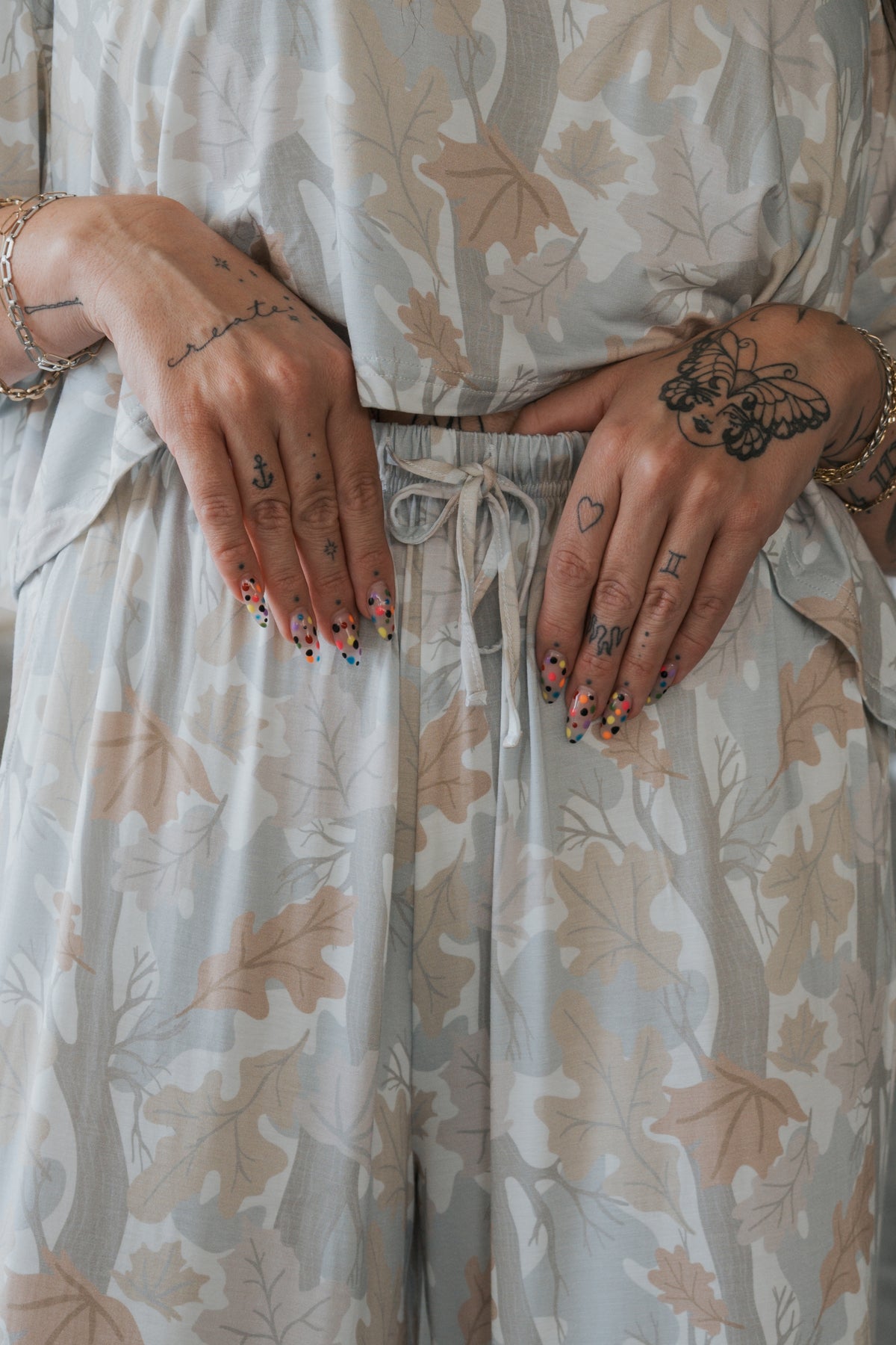 A person stands with hands on their waist, showing off colorful dotted nails and assorted tattoos—including a butterfly, an anchor, and hearts on their hands—while wearing the forever french baby Women’s Bamboo Crop Tee Lounge Set in Woodland. The leaf-patterned set features a light crop tee and wide-leg pants crafted from breathable bamboo fabric, contributing to a fresh and vibrant style.