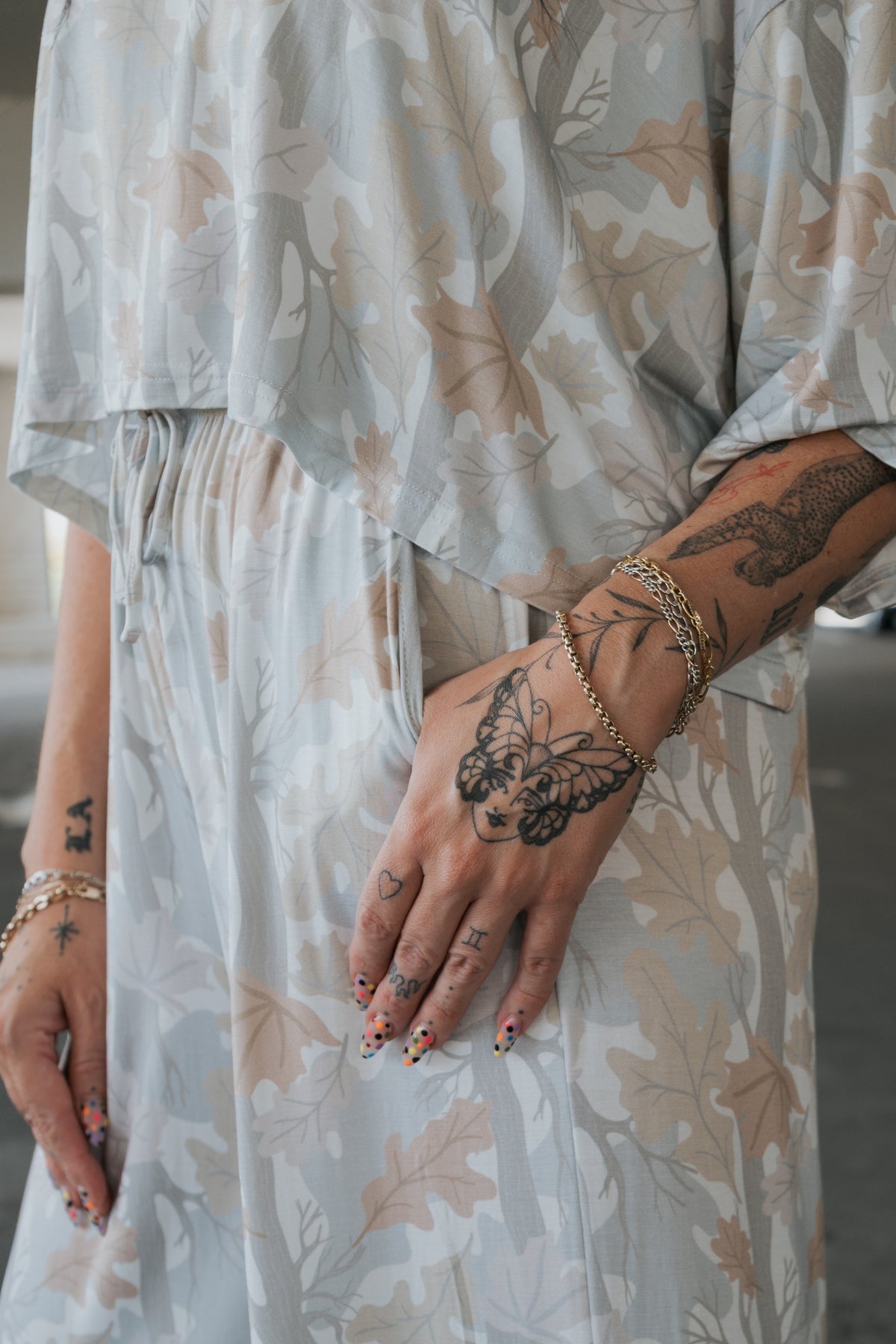 Wearing the forever french baby Women’s Bamboo Crop Tee Lounge Set in Woodland, a person showcases their tattooed hands and wrists adorned with gold bracelets. Their nails, decorated with a colorful design, draw attention—especially to the hand featuring a large butterfly tattoo.