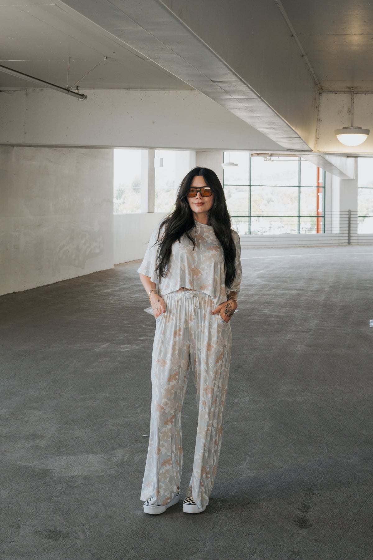 A woman with long dark hair and sunglasses stands in a spacious, empty parking garage, wearing the forever french baby Women’s Bamboo Crop Tee Lounge Set in the Woodland print.