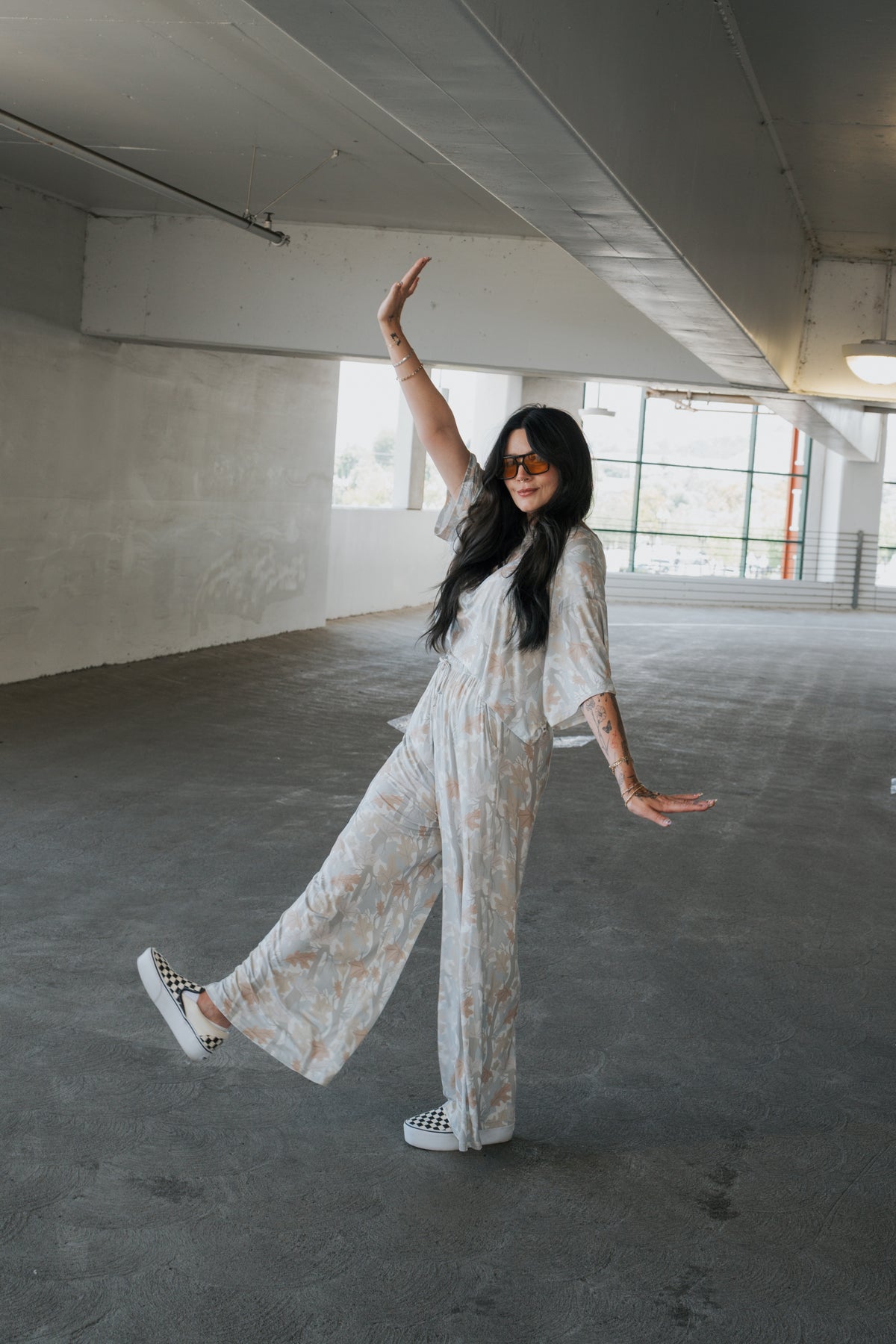 Wearing the Women’s Bamboo Crop Tee Lounge Set in Woodland by forever french baby, a woman with long dark hair, sunglasses, and visible tattoos playfully poses in a parking garage—raising one arm and balancing on one leg. She completes her look with checkered sneakers.