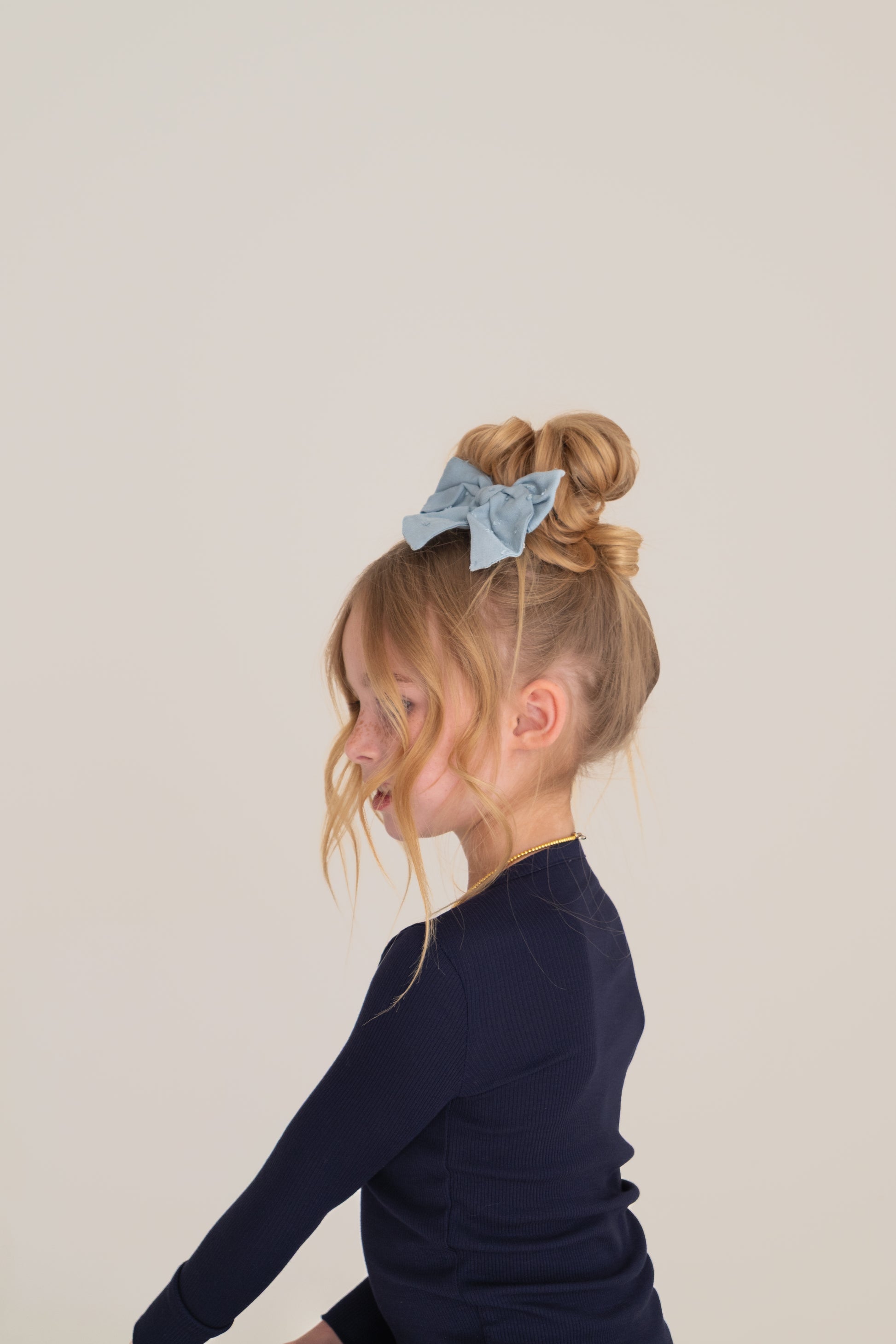 A young girl with blonde hair in a high bun and blue bow smiles sideways, wearing forever french baby Bamboo Two Piece Pajamas | Blueberry Ribbed, against a plain light background.