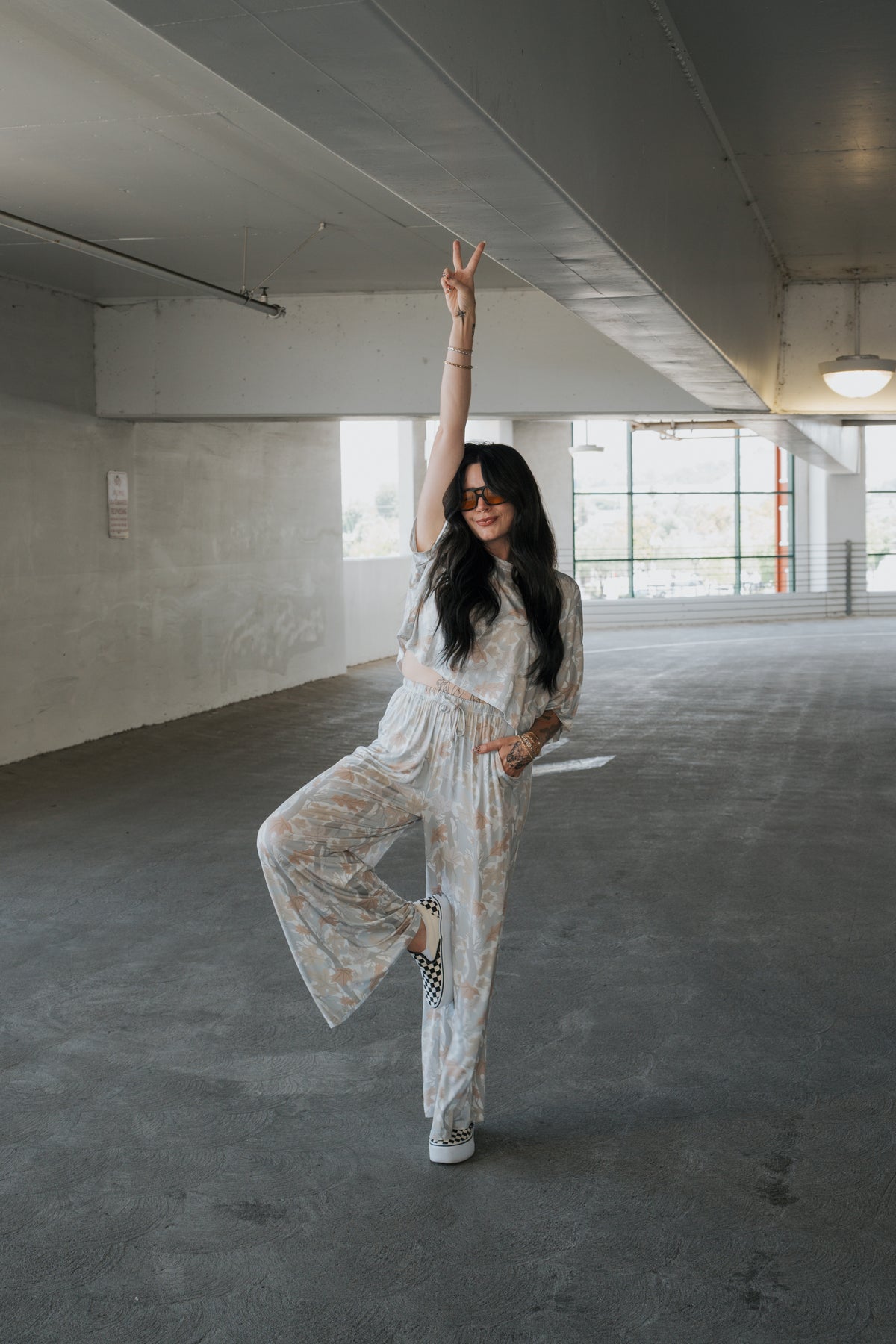 A woman, smiling and wearing sunglasses, poses on one leg in a parking garage, flashing a peace sign while dressed in the forever french baby Women’s Bamboo Crop Tee Lounge Set in Woodland.