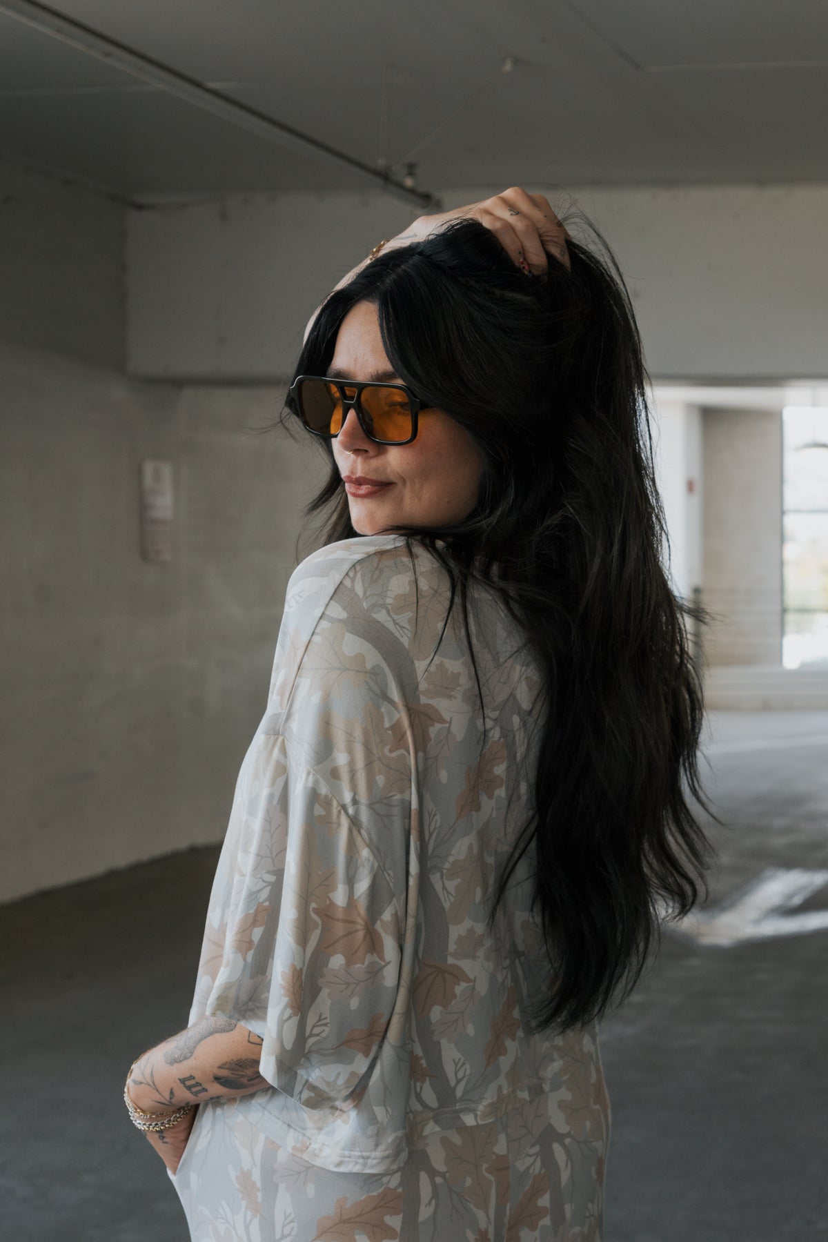 A woman with long dark hair and sunglasses stands in a parking garage, smiling slightly over her shoulder while wearing the forever french baby Women’s Bamboo Crop Tee Lounge Set in Woodland.