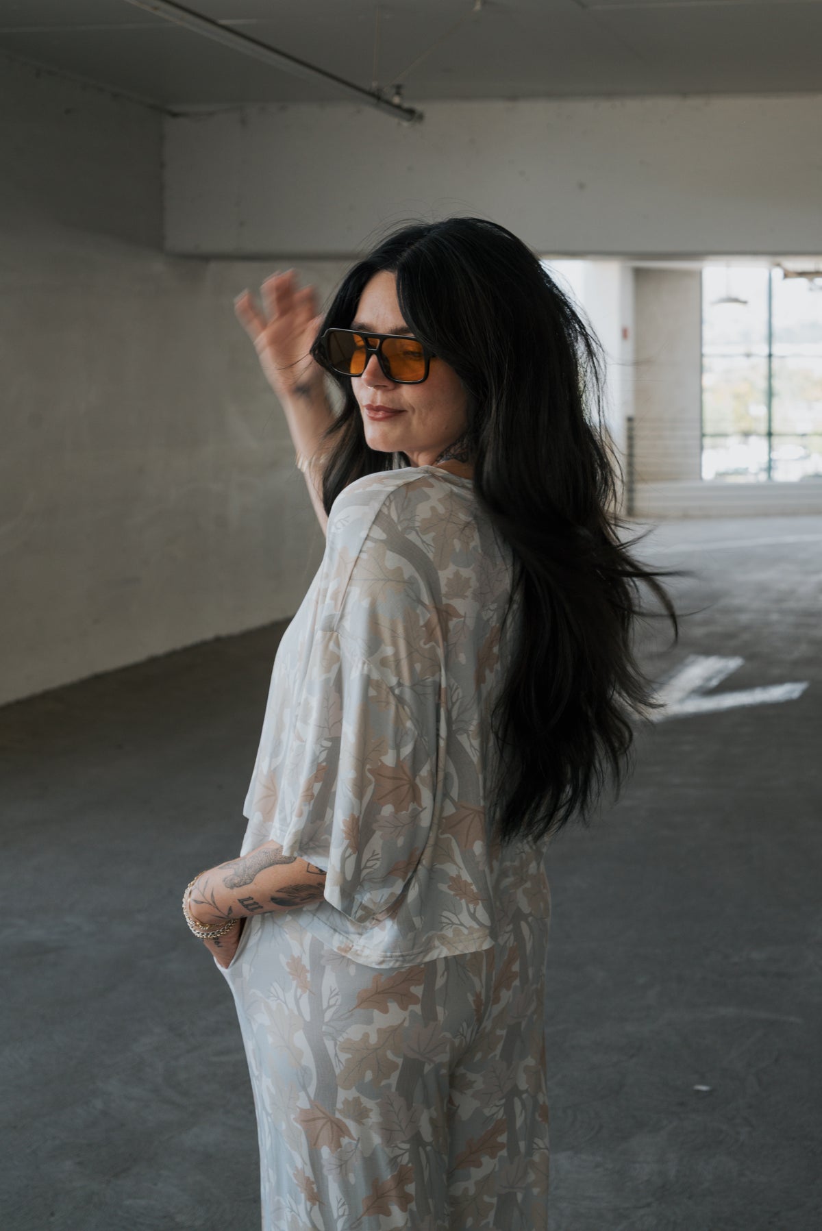 A woman with long dark hair wearing sunglasses and the forever french baby Women’s Bamboo Crop Tee Lounge Set in Woodland stands in a spacious, sunlit indoor area, looking back and smiling with one hand raised near her head.