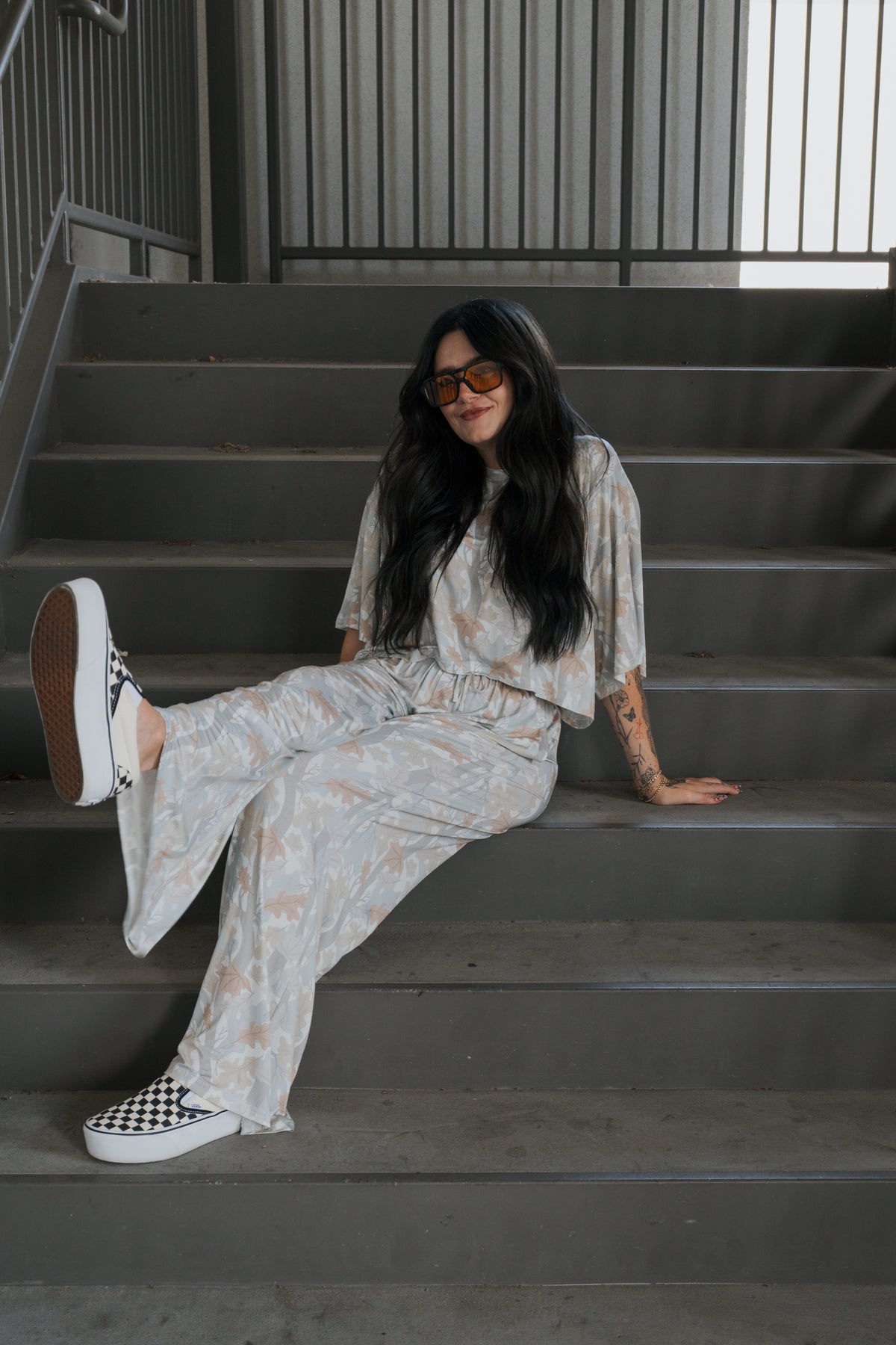 A woman with long dark hair, wearing sunglasses and the “Women’s Bamboo Crop Tee Lounge Set | Woodland” by forever french baby, sits on a staircase smiling, with one leg bent and her arm resting on a step. She completes her look with checkerboard slip-on shoes.