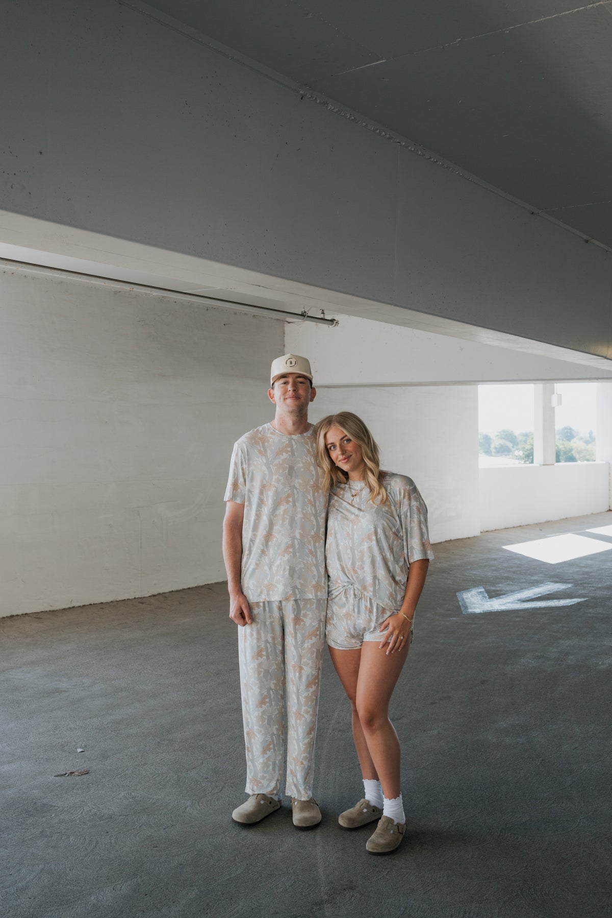 A man and woman stand together in a bright, empty parking garage, both wearing matching light-colored forever french baby loungewear. The man is dressed in the Men’s Bamboo Short Sleeve Pajama | Woodland, and the woman rests her head on his shoulder.