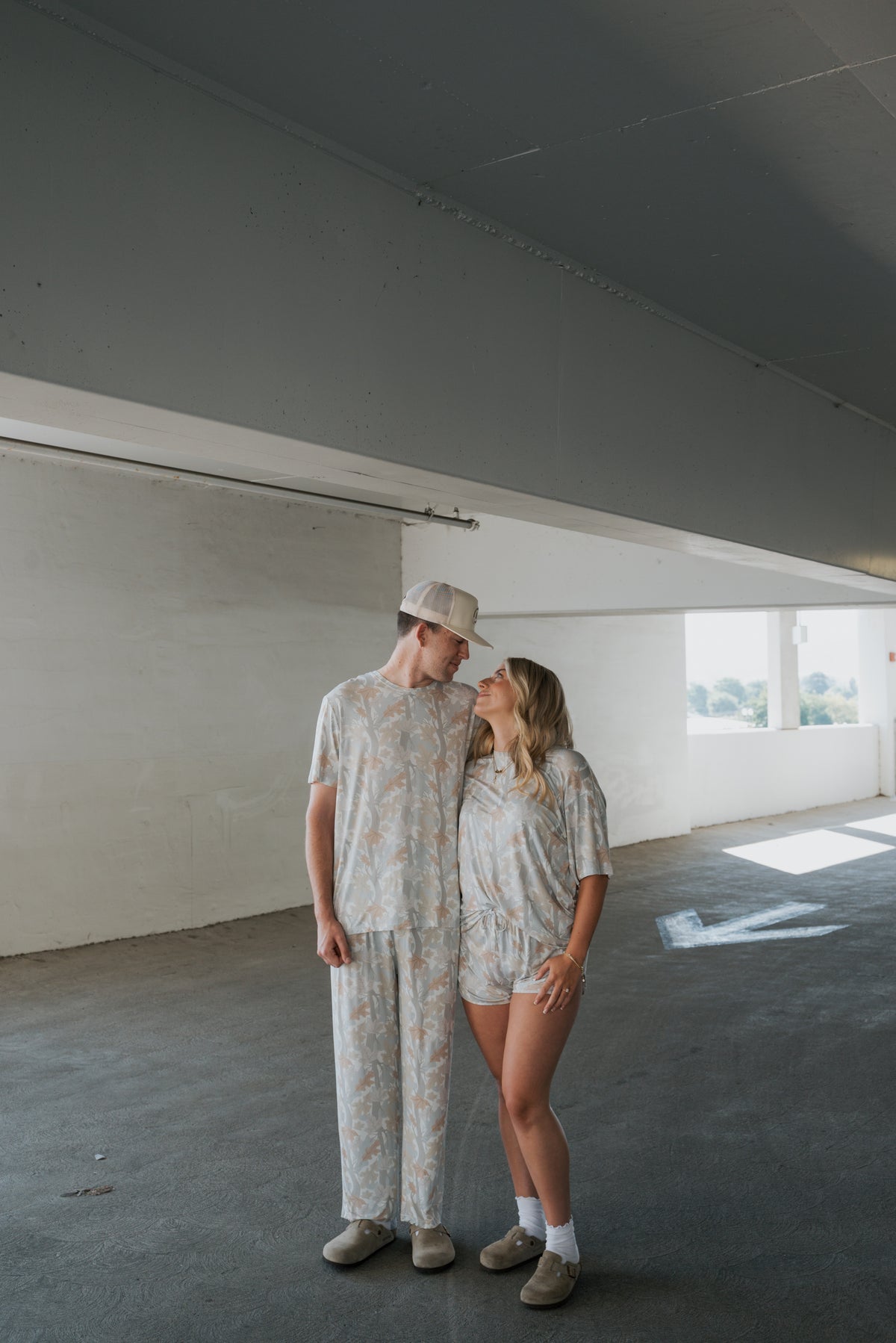 A couple stands in a parking garage, wearing matching light-colored patterned outfits from forever french baby. The man is dressed in the Men’s Bamboo Short Sleeve Pajama in Woodland, and both are facing each other and smiling as sunlight floods the space, emphasizing the softness and comfort of their bamboo clothing.