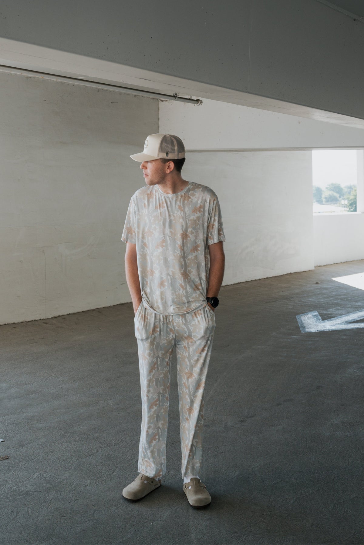 A person wearing the forever french baby Men’s Bamboo Short Sleeve Pajama in Woodland, along with a matching cap and slippers, stands in a concrete parking garage with hands in pockets, looking to the side as light streams through windows in the background.