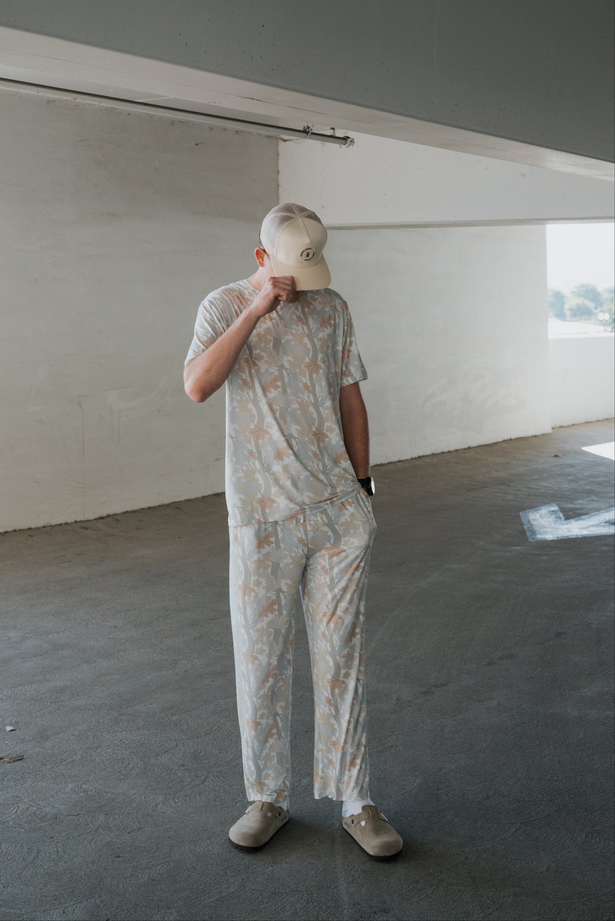 A person stands in a concrete parking garage, looking down and touching the brim of their light-colored cap while wearing the forever french baby Men’s Bamboo Short Sleeve Pajama in Woodland.