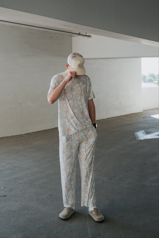 A person stands in a concrete parking garage, looking down and touching the brim of their light-colored cap while wearing the forever french baby Men’s Bamboo Short Sleeve Pajama in Woodland.