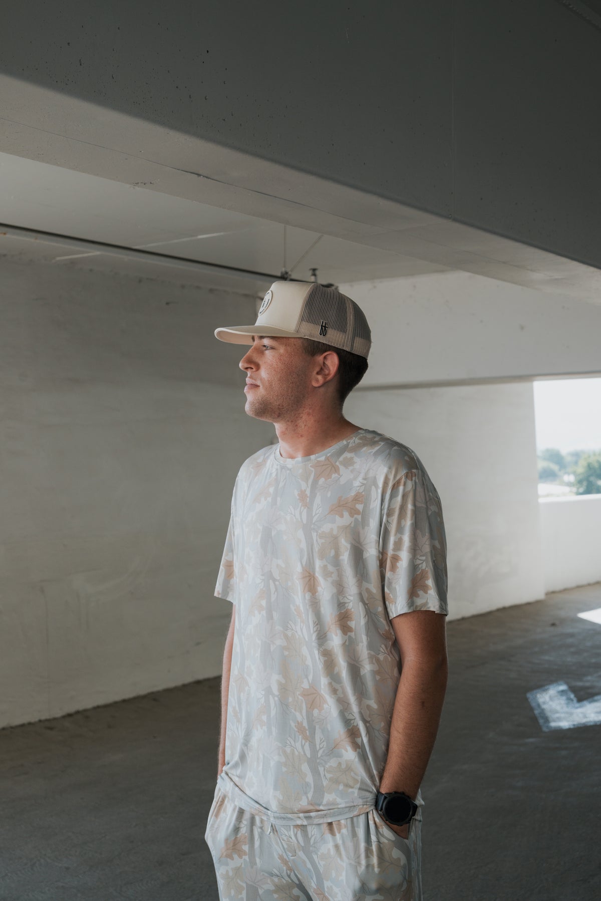 A man stands in a parking garage, looking to the side with his hands in his pockets. He is dressed in the forever french baby Men’s Bamboo Short Sleeve Pajama Set in Woodland and wears a beige cap for a stylish, coordinated look.