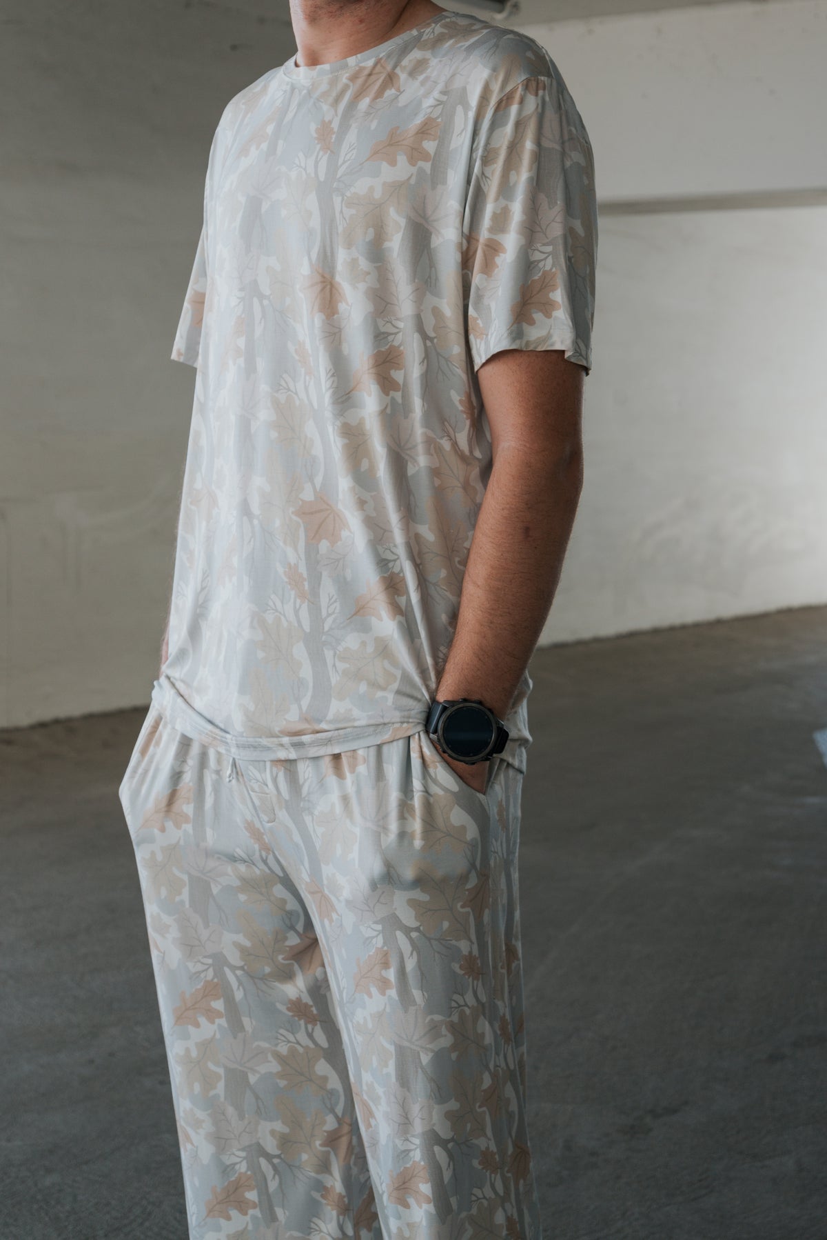 A person wearing the forever french baby Men’s Bamboo Short Sleeve Pajama in the Woodland pattern—featuring a subtle leaf design—stands in a concrete indoor space with one hand in a pocket and a black smartwatch on their wrist.