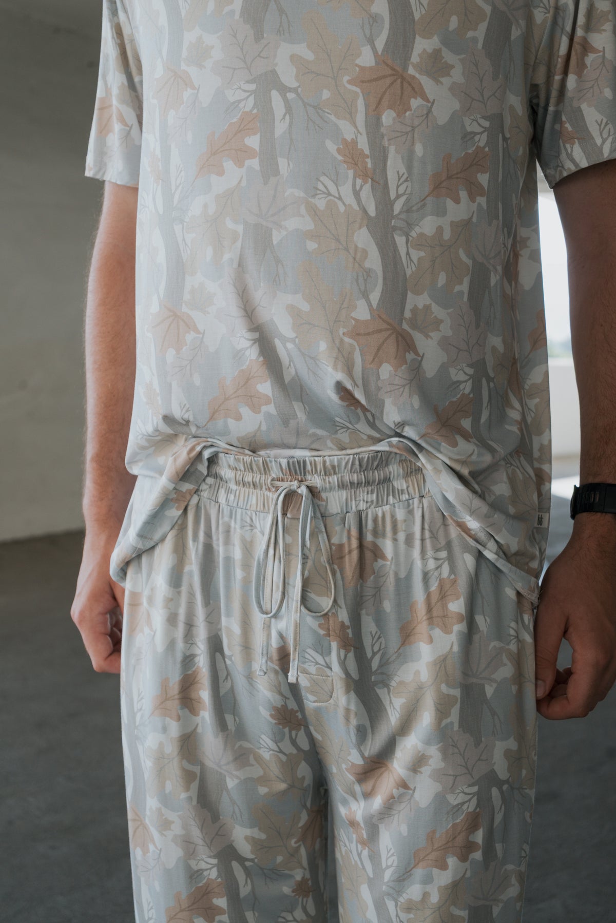 A person wearing the forever french baby Men’s Bamboo Short Sleeve Pajama in the Woodland print—with a matching short-sleeve shirt and drawstring pants featuring a light-colored leafy pattern—stands indoors, only their torso and arms visible.