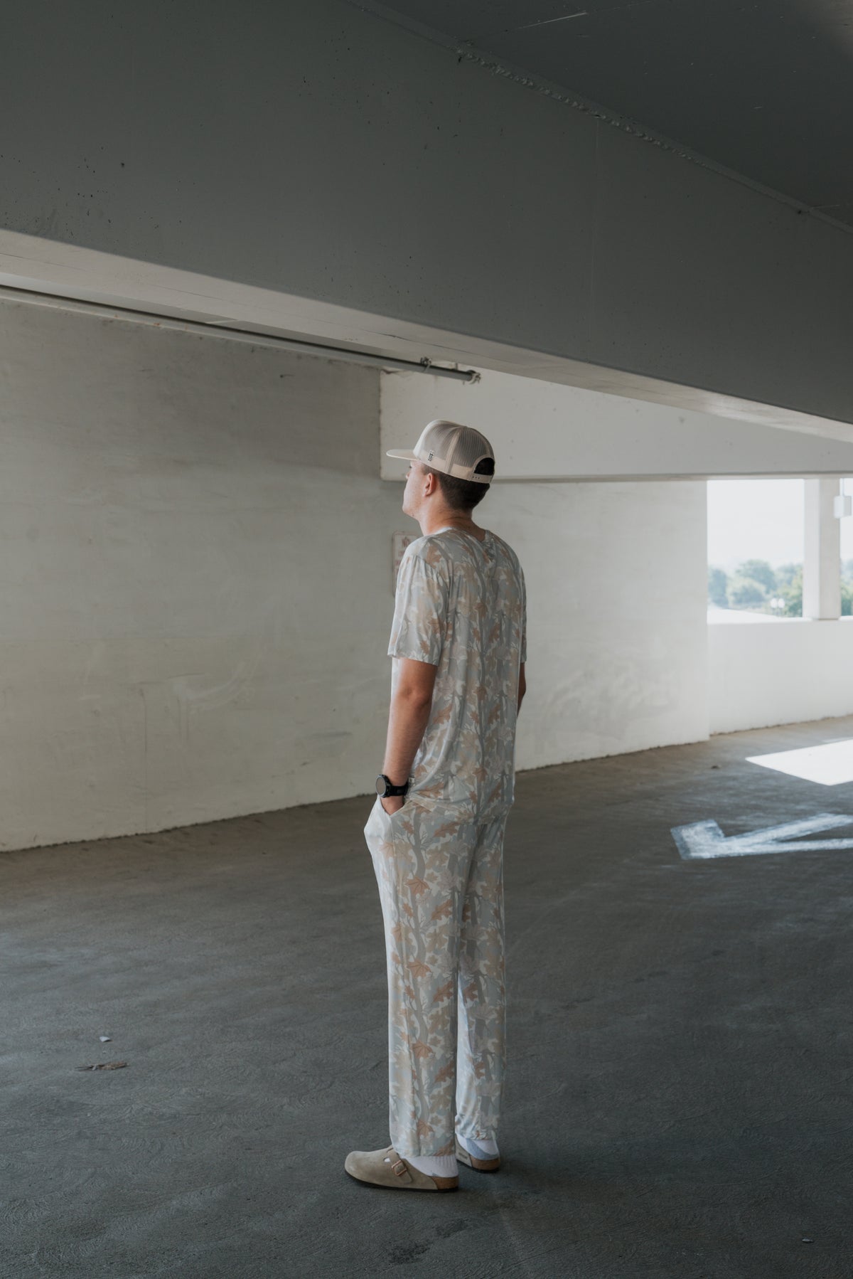 Wearing the forever french baby Men’s Bamboo Short Sleeve Pajama in the Woodland print, a person stands in a mostly empty concrete parking garage with hands in pockets, facing a wall as sunlight streams through large openings behind them.