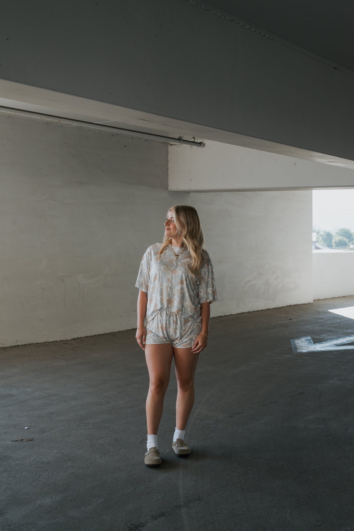 A woman with long blonde hair stands in a concrete parking garage, wearing the Women’s Bamboo Short Set in Woodland by forever french baby, paired with white socks and sneakers. She gazes to the side as sunlight streams through an opening behind her.