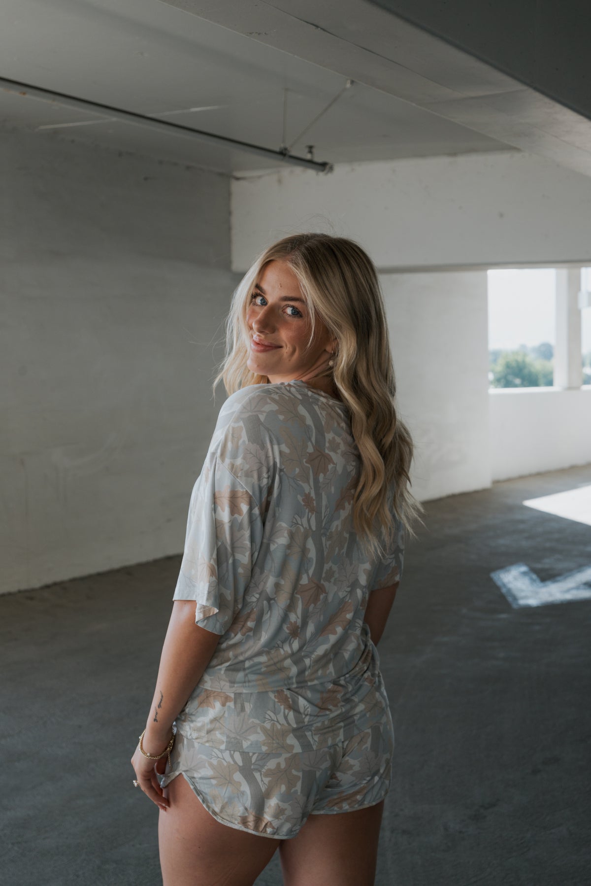 Wearing the “Women’s Bamboo Short Set | Woodland” by forever french baby, a woman with long blonde hair stands in a sunlit parking garage, looking back over her shoulder and smiling.