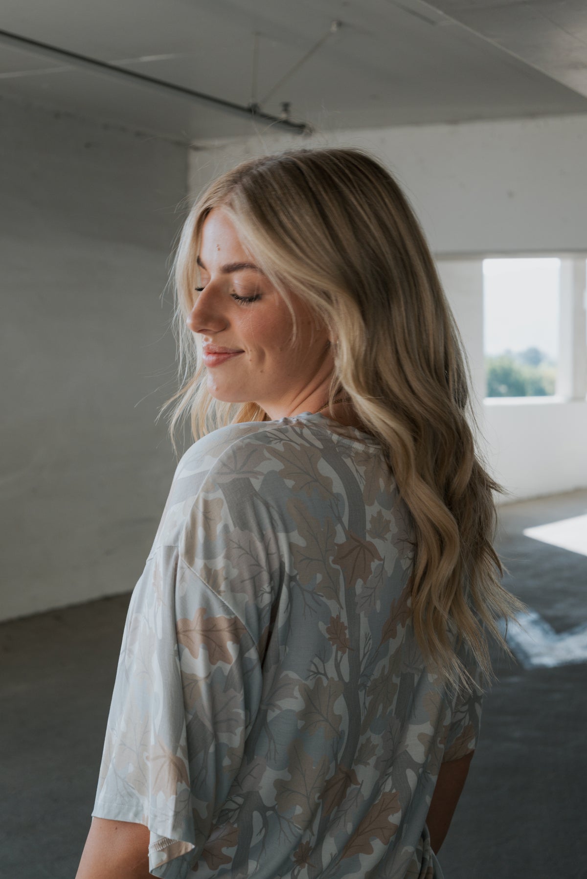 A woman with long blonde hair stands indoors, smiling gently with her eyes closed as sunlight streams through the windows behind her. She is wearing the Women’s Bamboo Short Set in Woodland by forever french baby, made from soft, hypo-allergenic bamboo fabric.