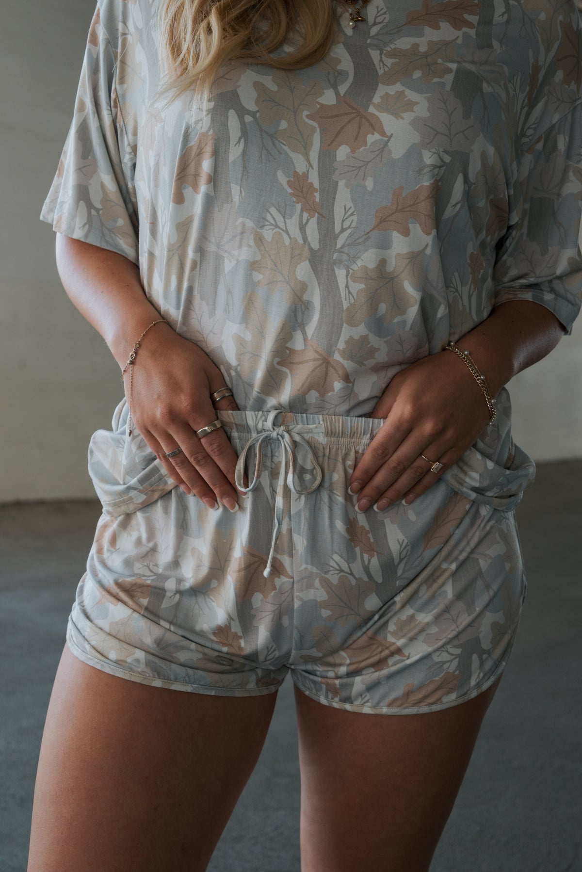 A person is shown from the torso to upper legs wearing the forever french baby Women’s Bamboo Short Set | Woodland—a light-colored, leaf-patterned matching short-sleeve top and shorts crafted from hypo-allergenic bamboo—with hands resting in the shorts pockets.