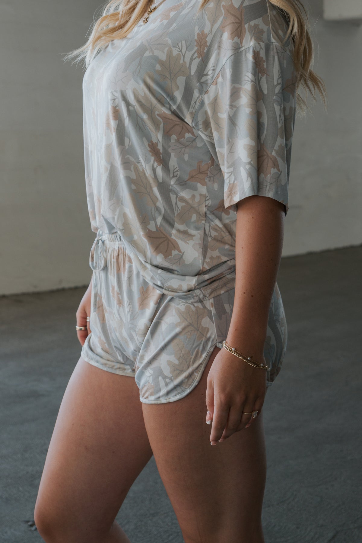 A person is standing indoors wearing the forever french baby Women’s Bamboo Short Set in the Woodland print, a light-colored, floral-patterned short-sleeve top and matching shorts made from soft, hypo-allergenic bamboo. Their face is not visible.