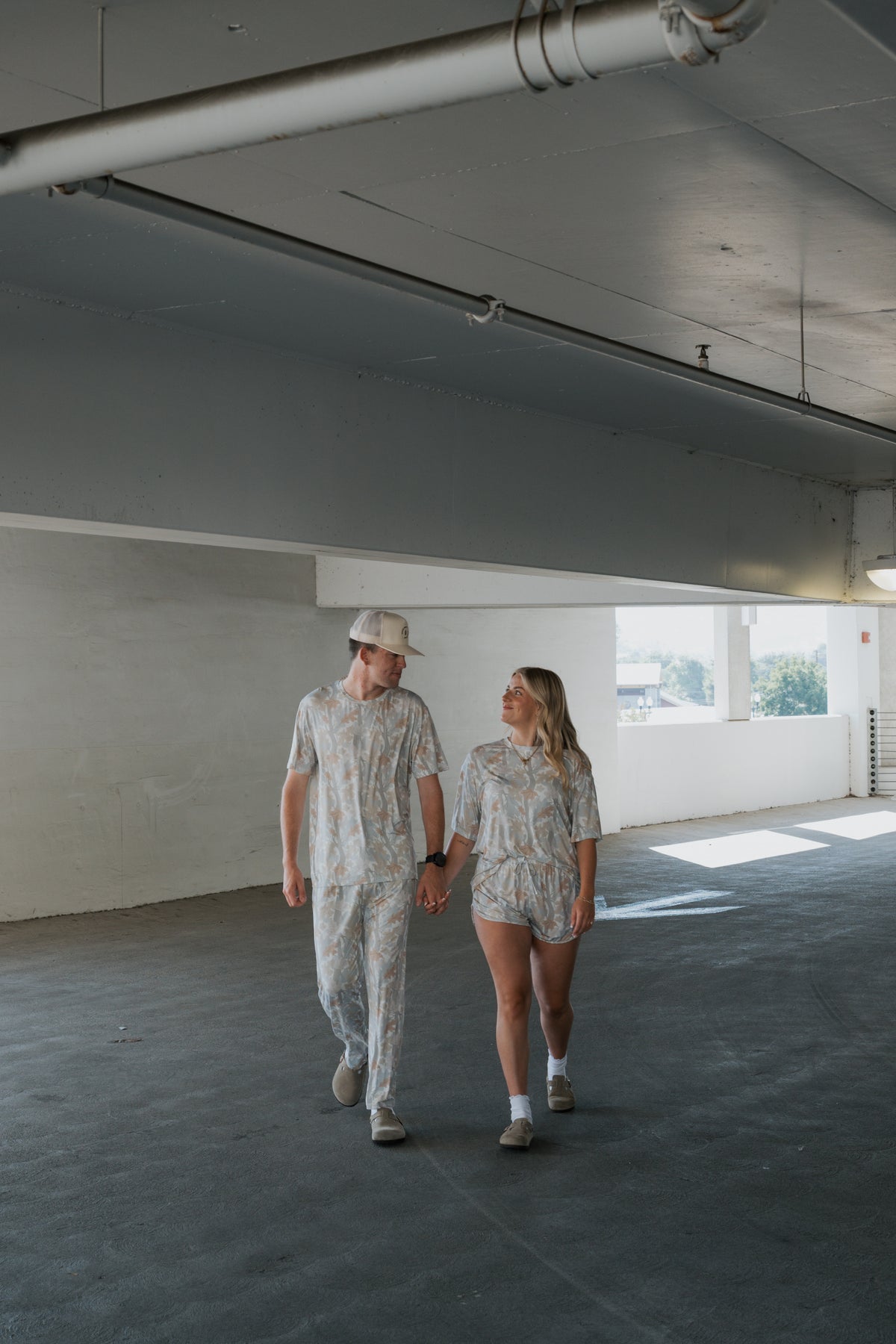 A couple, with the woman wearing the forever french baby Women’s Bamboo Short Set in Woodland, walks hand in hand through a bright, empty parking garage, looking at each other and smiling.
