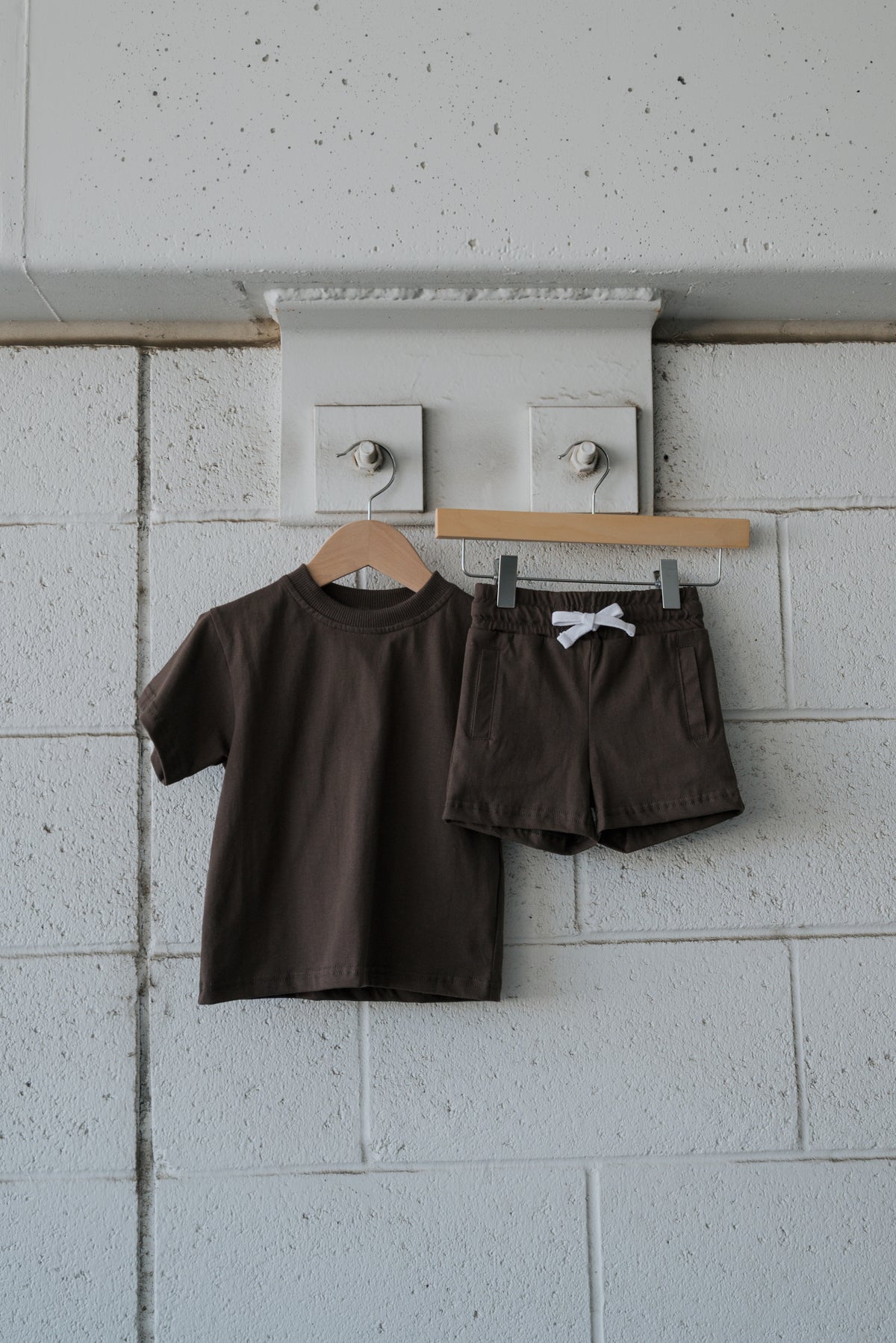 The Child Short Set | Espresso by forever french baby, a brown short-sleeve tee and matching shorts made from 100% cotton with a white drawstring, is displayed on a wooden hanger against a white textured wall.