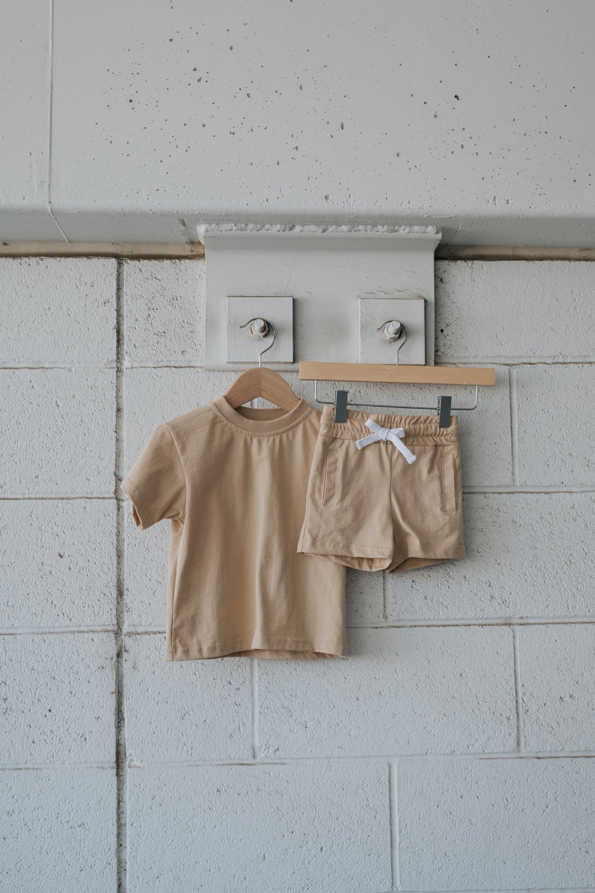 A Child Short Set | Toffee by forever french baby—a beige cotton ensemble comprising a short-sleeve t-shirt and matching shorts—hangs on wooden hangers against a white-painted brick wall accented with large bolts and topped by a concrete ledge. Ideal for everyday wear, this comfortable set offers both style and practicality, making it the perfect play outfit for active children.