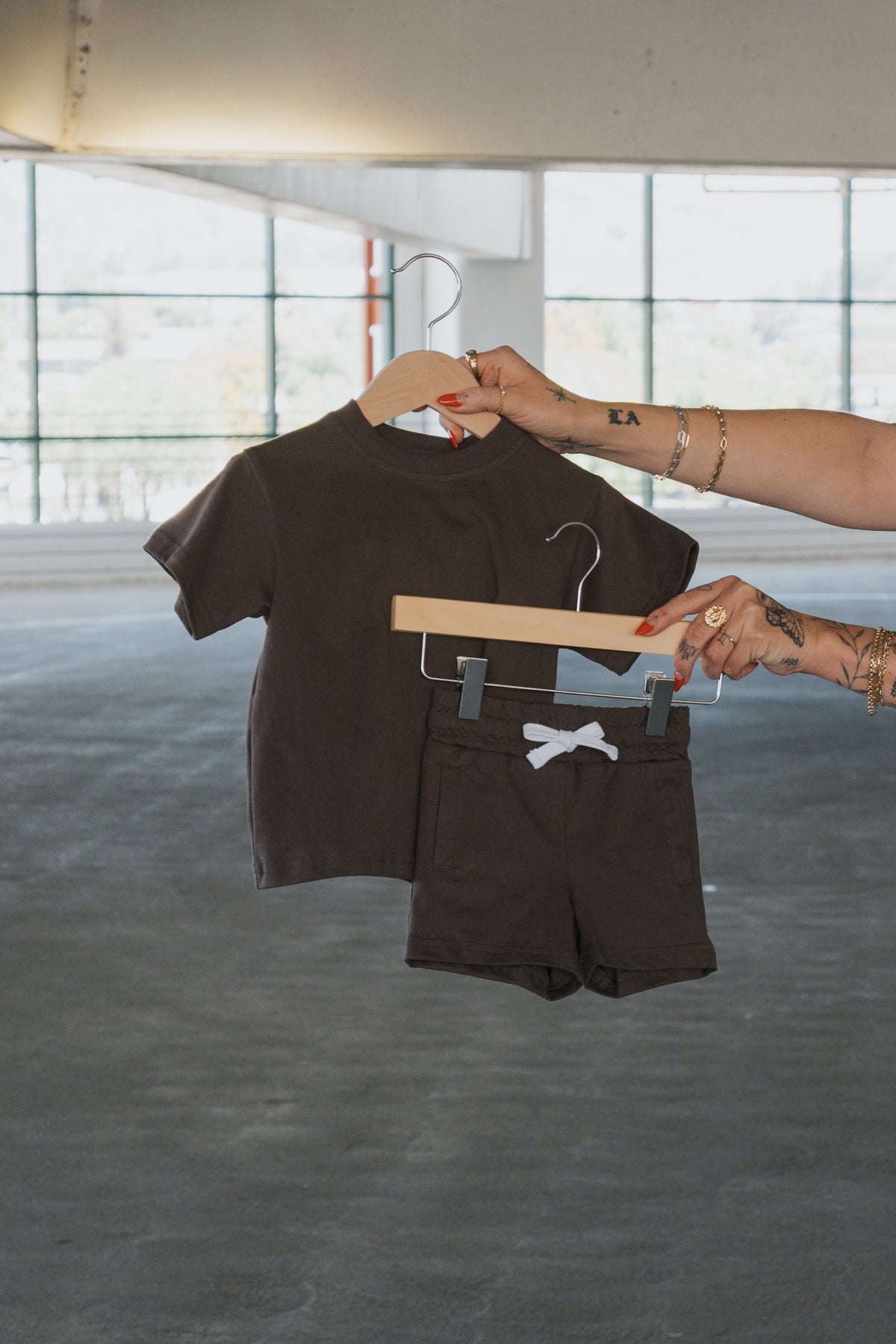 A person holds up the forever french baby Child Short Set in Espresso—made from 100% cotton, featuring a matching short-sleeve tee and kids shorts with a white drawstring—on separate hangers in a bright, empty parking garage.
