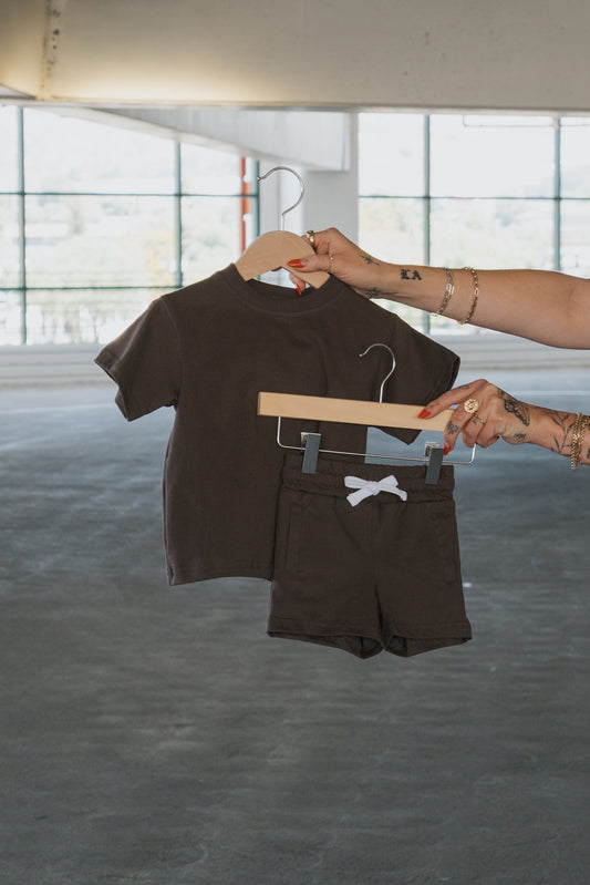 A person holds up the forever french baby Child Short Set in Espresso—made from 100% cotton, featuring a matching short-sleeve tee and kids shorts with a white drawstring—on separate hangers in a bright, empty parking garage.