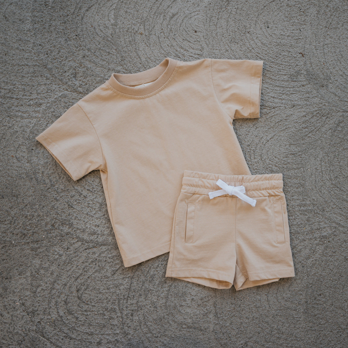The forever french baby Child Short Set | Toffee features a beige short-sleeve t-shirt and matching kids cotton shorts with a white drawstring, laid out flat on a textured gray surface—ideal as comfortable play clothes and everyday wear for children.