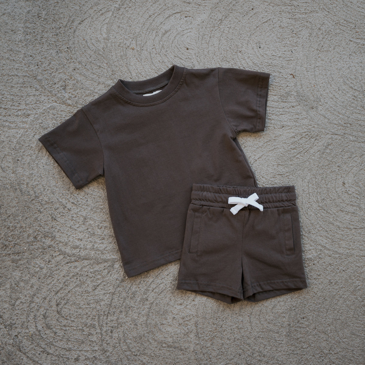 The forever french baby Child Short Set | Espresso features a matching dark brown toddler ensemble made from 100% cotton, including a short-sleeve t-shirt and shorts adorned with a white bow, displayed on a textured concrete surface—perfect for everyday comfort and style.