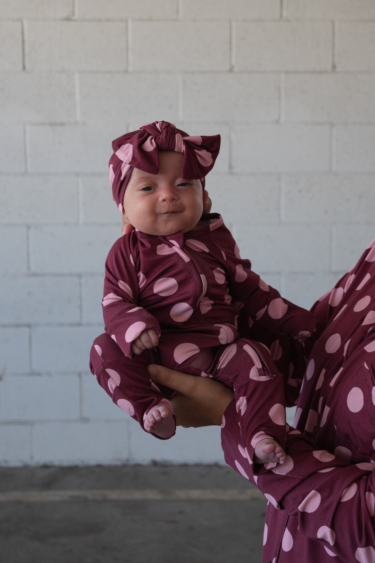 A baby wearing a coordinated maroon ensemble with large pink polka dots and the Forever French Baby Bamboo Head Wrap in Heather Rose Dot is being held up by an adult in front of a white brick wall, perfectly capturing the signature Forever French Baby style.