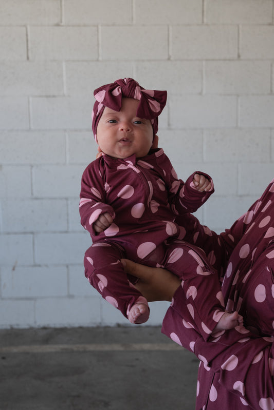 A baby in the forever french baby Bamboo Zip Pajamas | Heather Rose Dot—featuring a maroon design with large pink polka dots and a matching headband—is held by an adult against a white brick wall, modeling adorable and breathable hypoallergenic sleepwear.