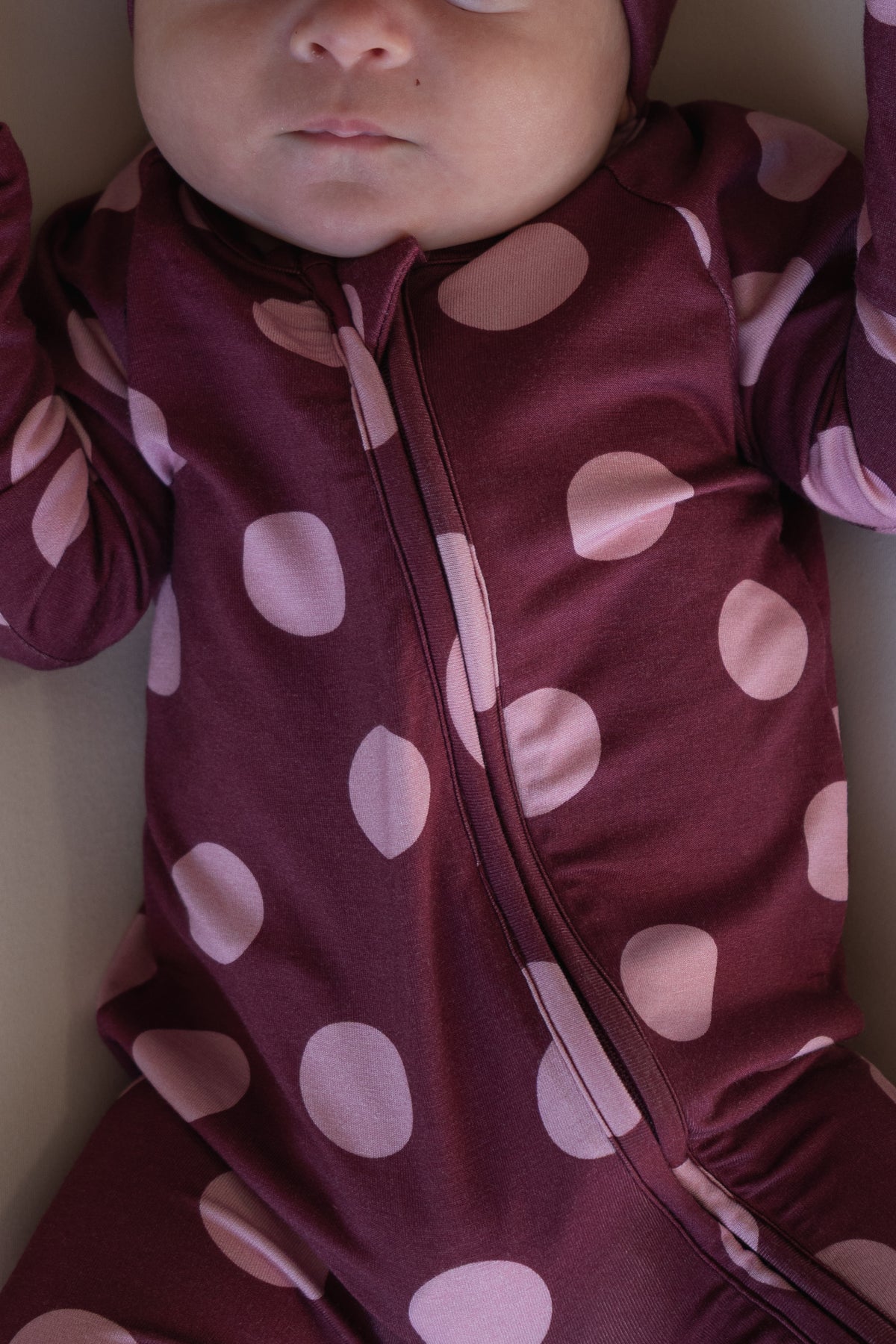 A baby wearing forever french baby’s Bamboo Zip Pajamas in Heather Rose Dot—a maroon outfit with large light pink polka dots—lies on their back, arms raised, with only the lower face visible.
