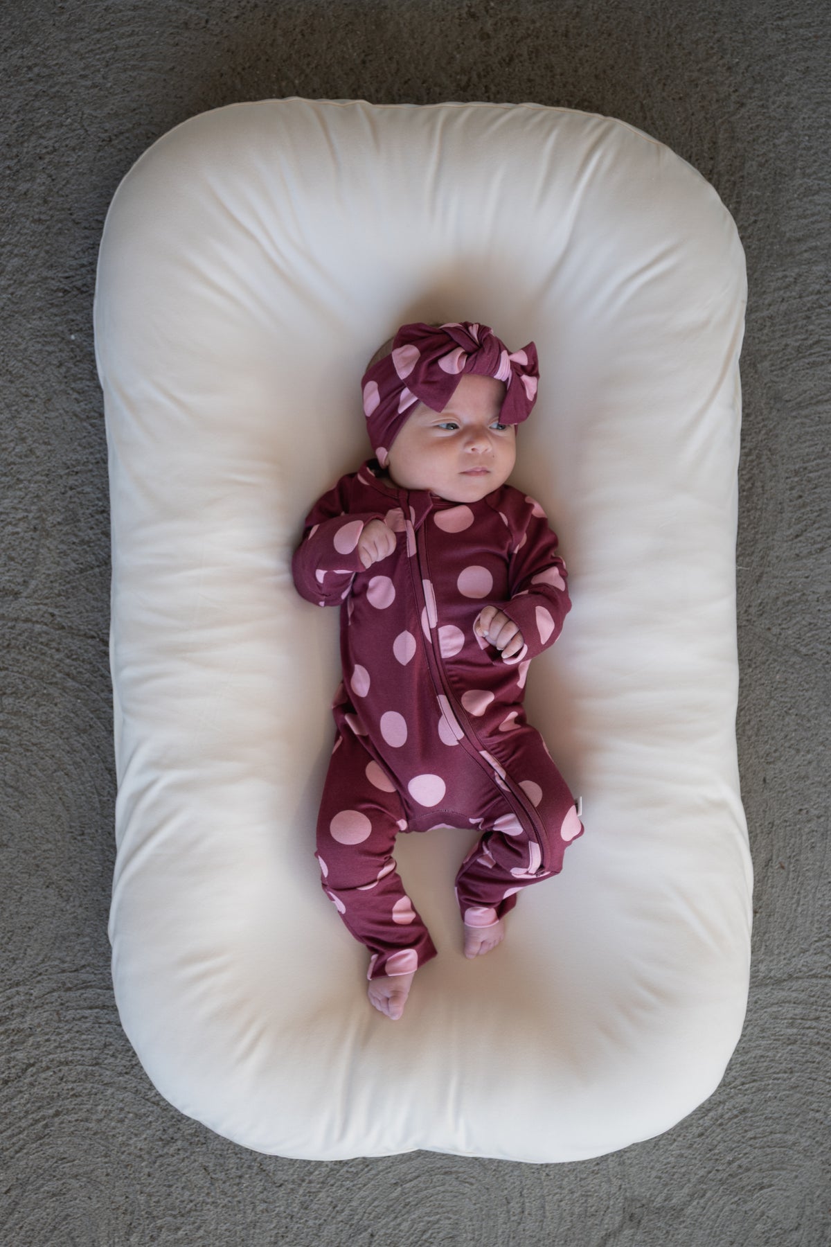 A baby wearing a maroon outfit with large pink polka dots and a matching Forever French Baby Bamboo Head Wrap in Heather Rose Dot lies on a white, oval-shaped cushion atop a gray textured surface.