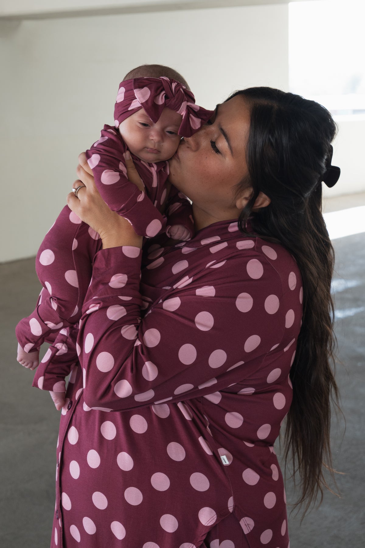 A woman in a maroon and pink polka dot outfit kisses a baby on the cheek. The baby, wearing forever french baby’s Bamboo Zip Pajamas in Heather Rose Dot with a matching headband, looks at the camera. They are indoors, where soft natural light highlights their cozy and breathable pajamas.