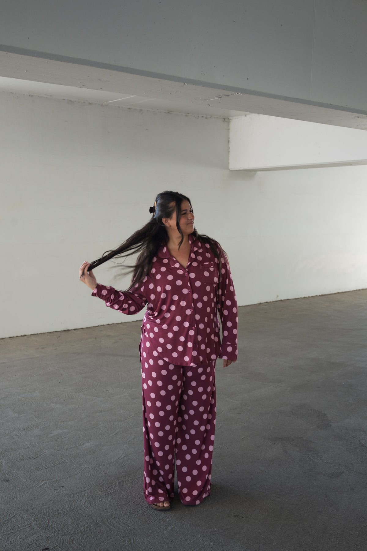 A person is standing indoors on a gray floor in front of a neutral white wall, wearing the forever french baby Women’s Bamboo Button Up Lounge Set in Heather Rose Dot—featuring white polka dots and wide leg lounge pants—while holding and looking at their long hair.