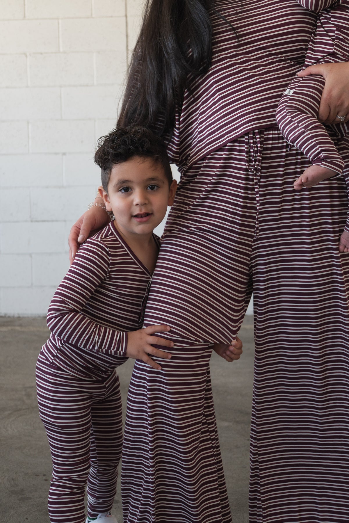 A young child in matching maroon striped pajamas from Forever French Baby hugs an adult’s leg, while the adult—also wearing Bamboo Two Piece Pajamas in Maroon Stripe—holds a baby. Only part of the adults’ faces are visible as they stand in front of a white brick wall, all dressed in soft, breathable kids sleepwear made from hypoallergenic bamboo fabric for ultimate comfort and peace of mind.