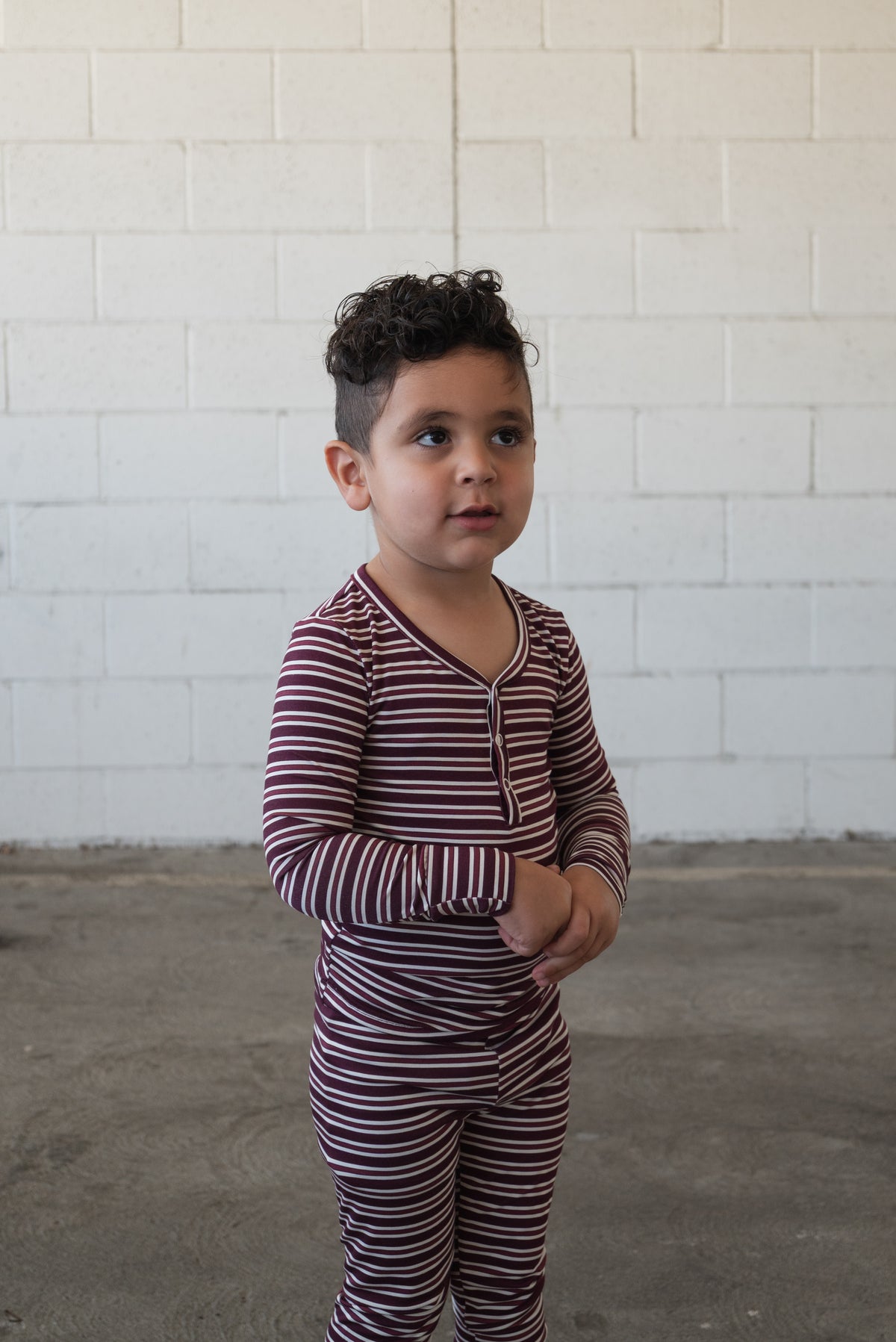A young child with curly hair stands indoors against a light-colored brick wall, wearing the forever french baby Bamboo Two Piece Pajamas in Maroon Stripe. This breathable kids’ sleepwear ensures comfort throughout the night.