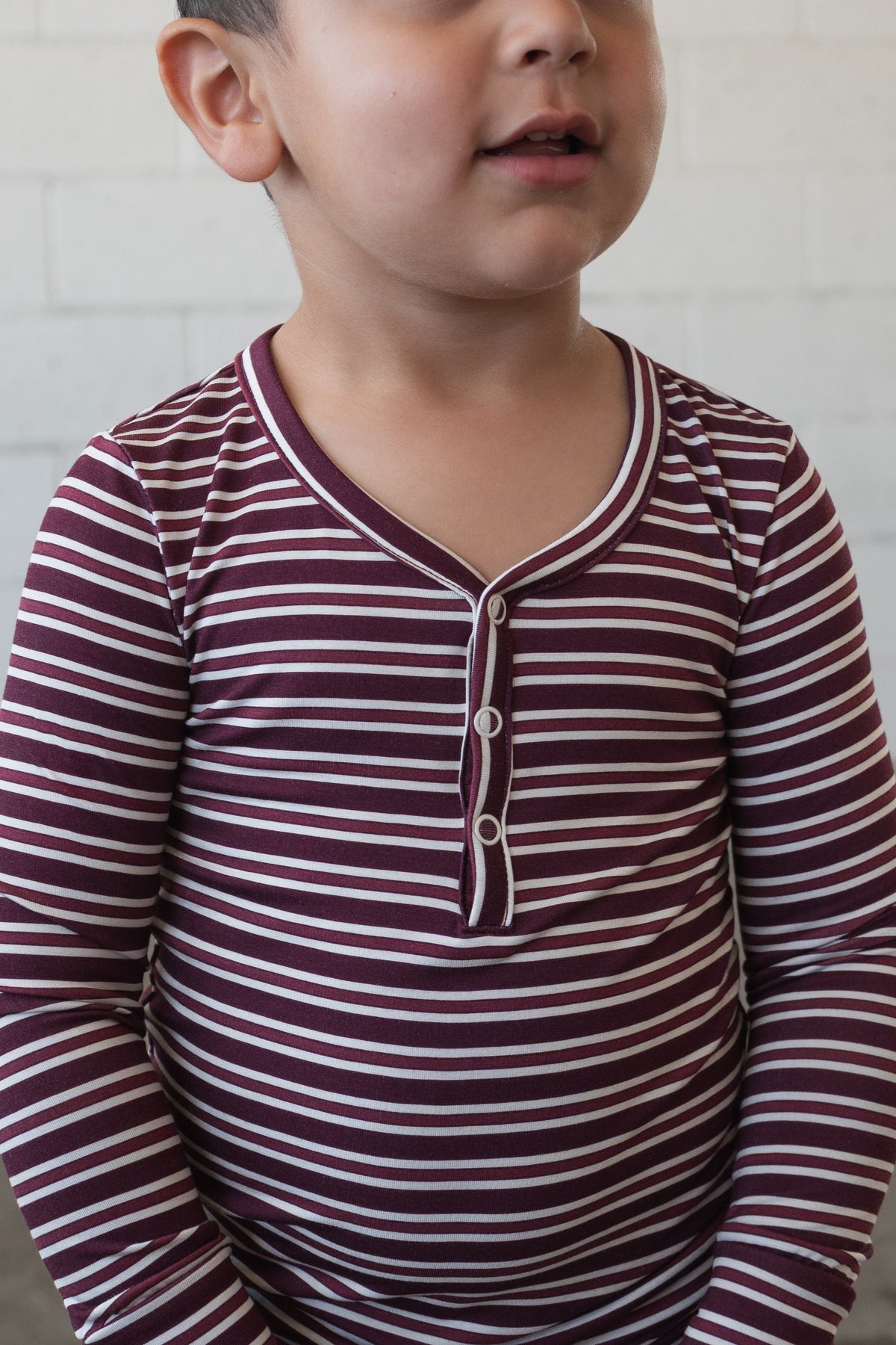 A young child, partially smiling, stands in front of a white brick wall wearing forever french baby’s Bamboo Two Piece Pajamas in Maroon Stripe, featuring maroon and white stripes with long sleeves and button details.