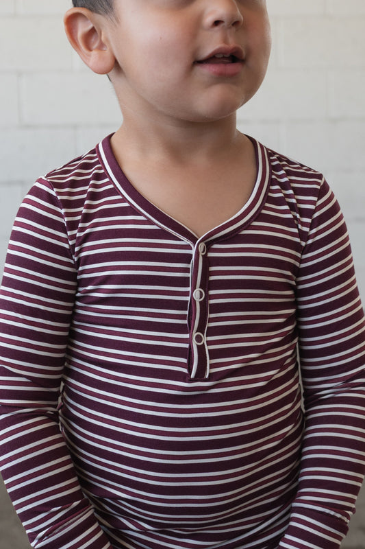 A young child, partially smiling, stands in front of a white brick wall wearing forever french baby’s Bamboo Two Piece Pajamas in Maroon Stripe, featuring maroon and white stripes with long sleeves and button details.