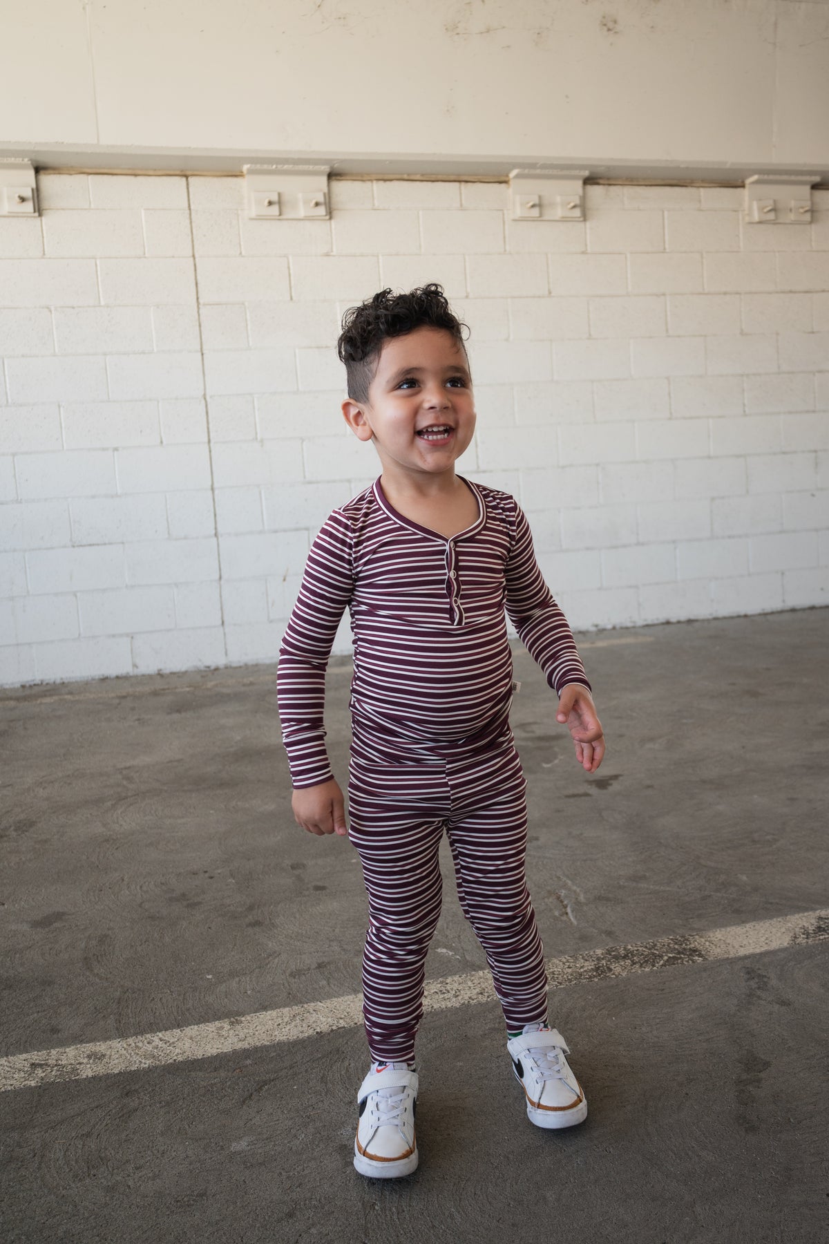 A young child wearing forever french baby’s Bamboo Two Piece Pajamas in Maroon Stripe stands smiling on a concrete floor in front of a white brick wall.