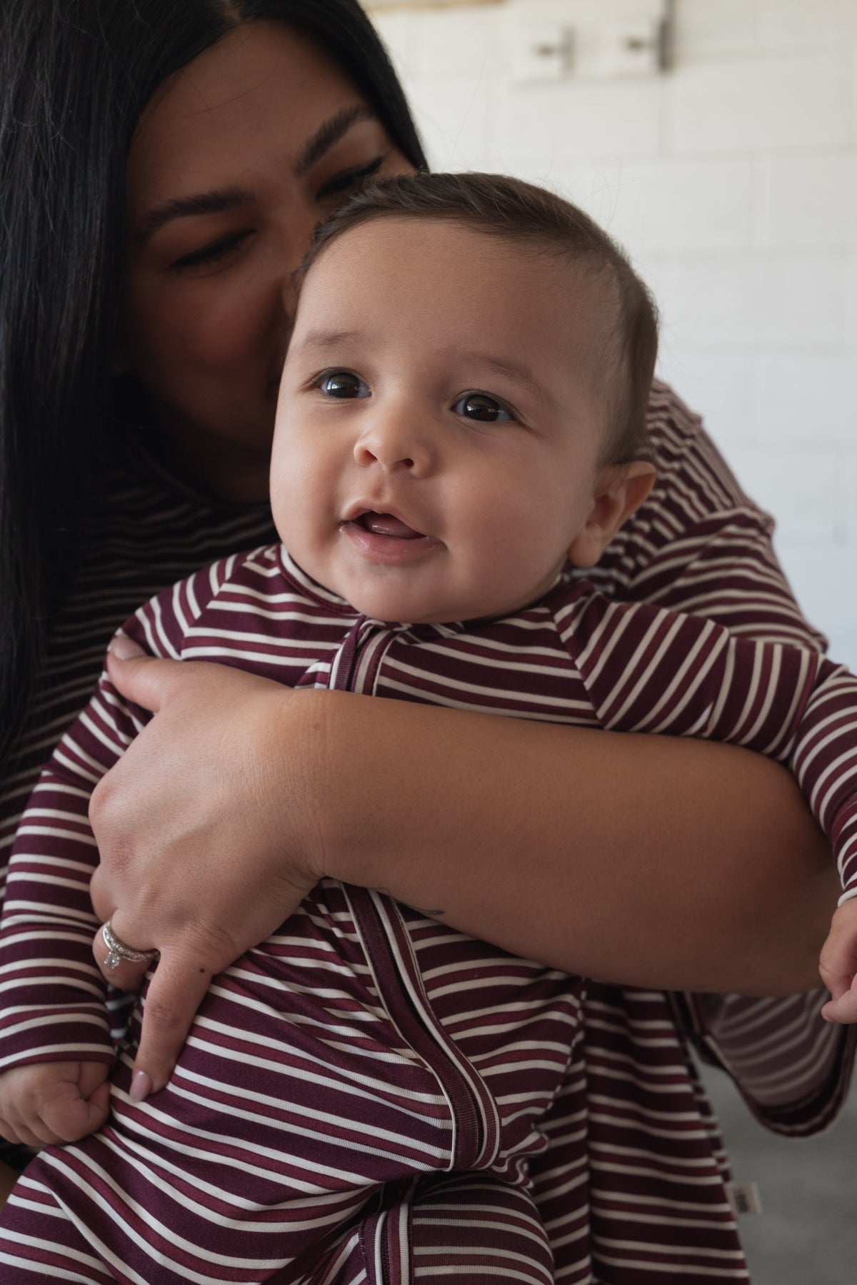 A woman hugs and kisses a smiling baby, both dressed in matching forever french baby Bamboo Zip Pajamas in Maroon Stripe. The breathable, hypoallergenic bamboo fabric ensures comfort for the little one as they look forward, while the woman holds them close.