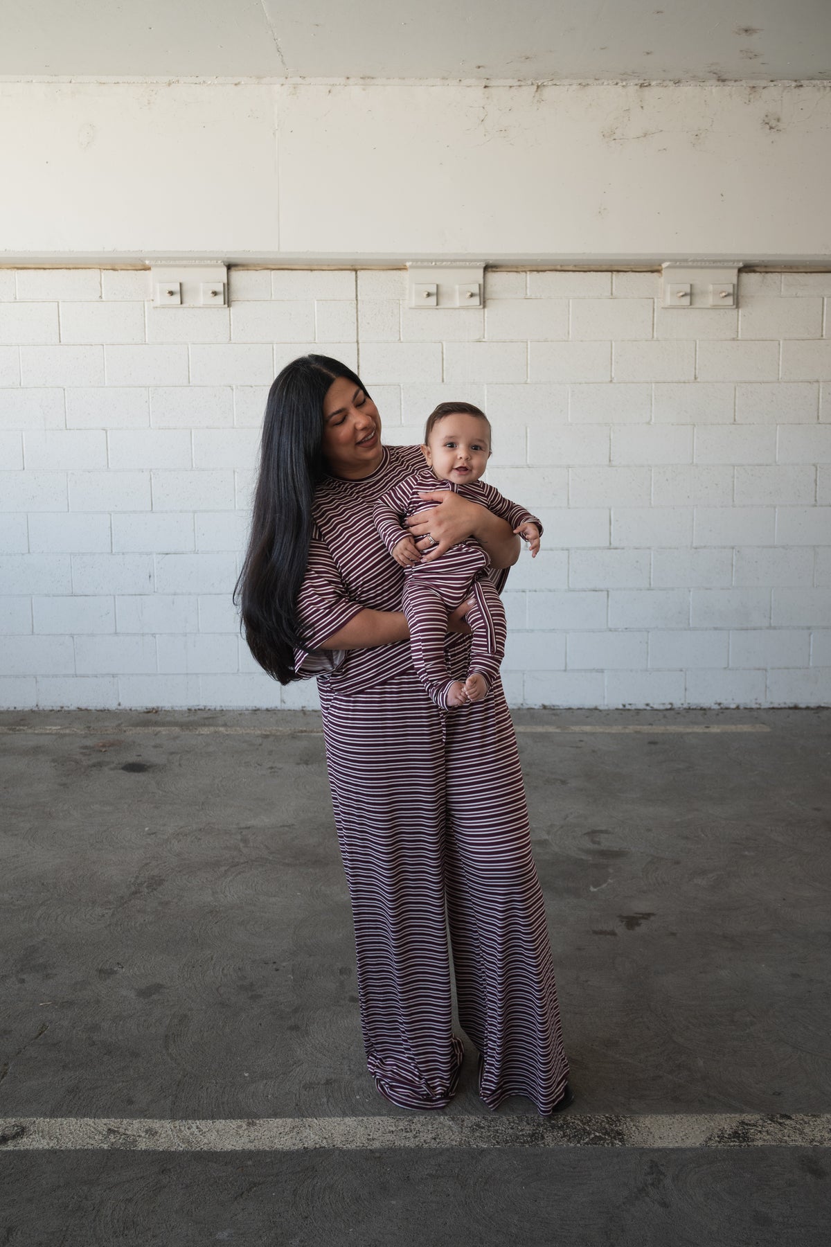 A woman and a baby, both dressed in matching Bamboo Zip Pajamas in Maroon Stripe by forever french baby, stand in a concrete parking area in front of a white brick wall. The woman is smiling while holding the baby.