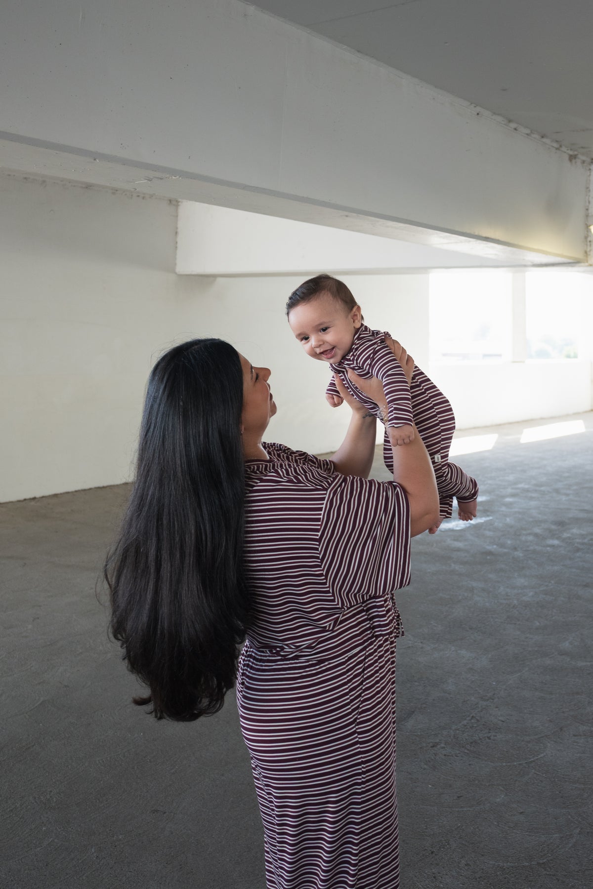 A woman with long dark hair in a striped dress lifts a smiling baby, dressed in forever french baby’s Bamboo Zip Pajamas in Maroon Stripe, in a bright and spacious indoor setting.
