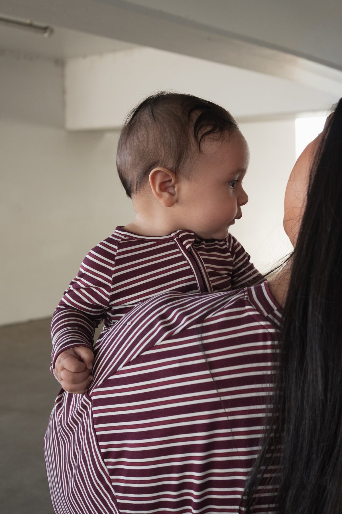 A baby wearing forever french baby’s Bamboo Zip Pajamas in Maroon Stripe is held over the shoulder of an adult dressed in a coordinating striped shirt, both turned away in a softly lit room.