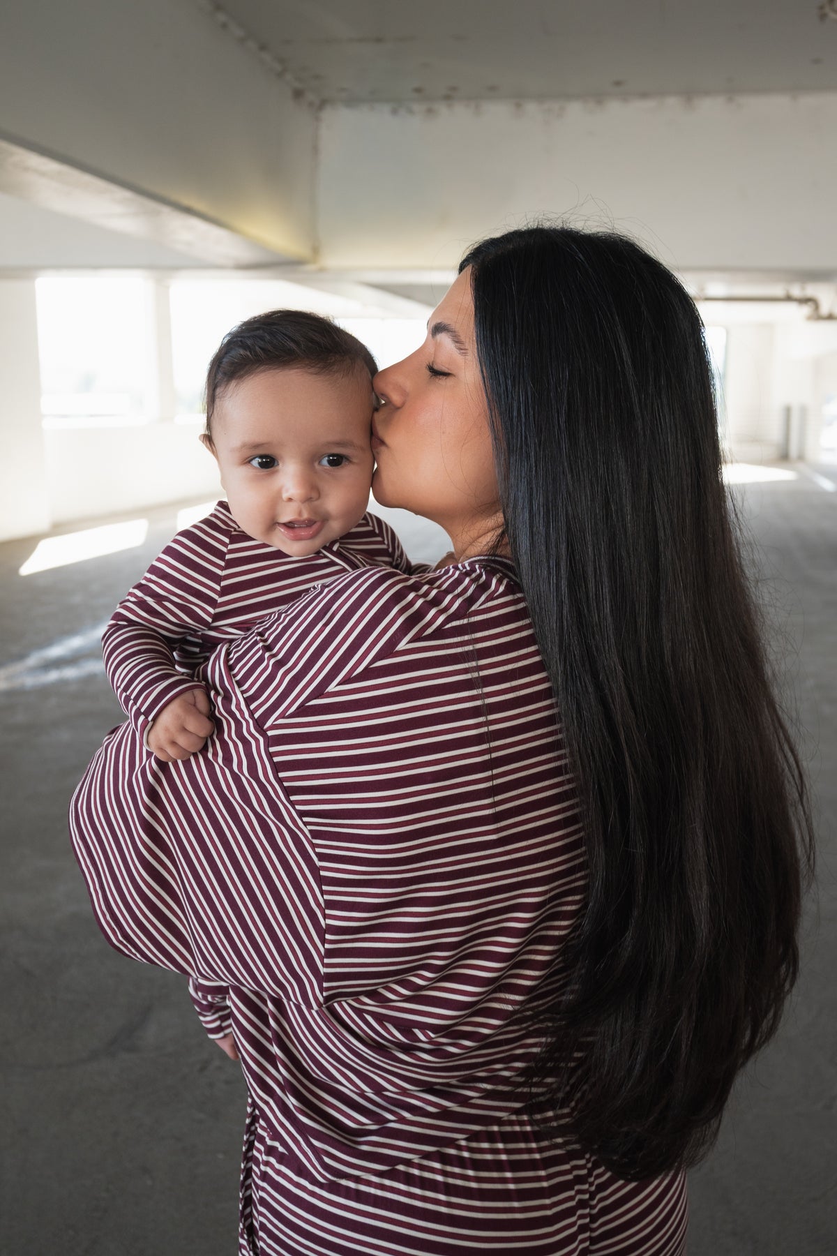 A woman with long dark hair kisses a baby on the cheek. Both are wearing matching forever french baby Bamboo Zip Pajamas in Maroon Stripe as they stand in a sunlit, indoor parking garage.